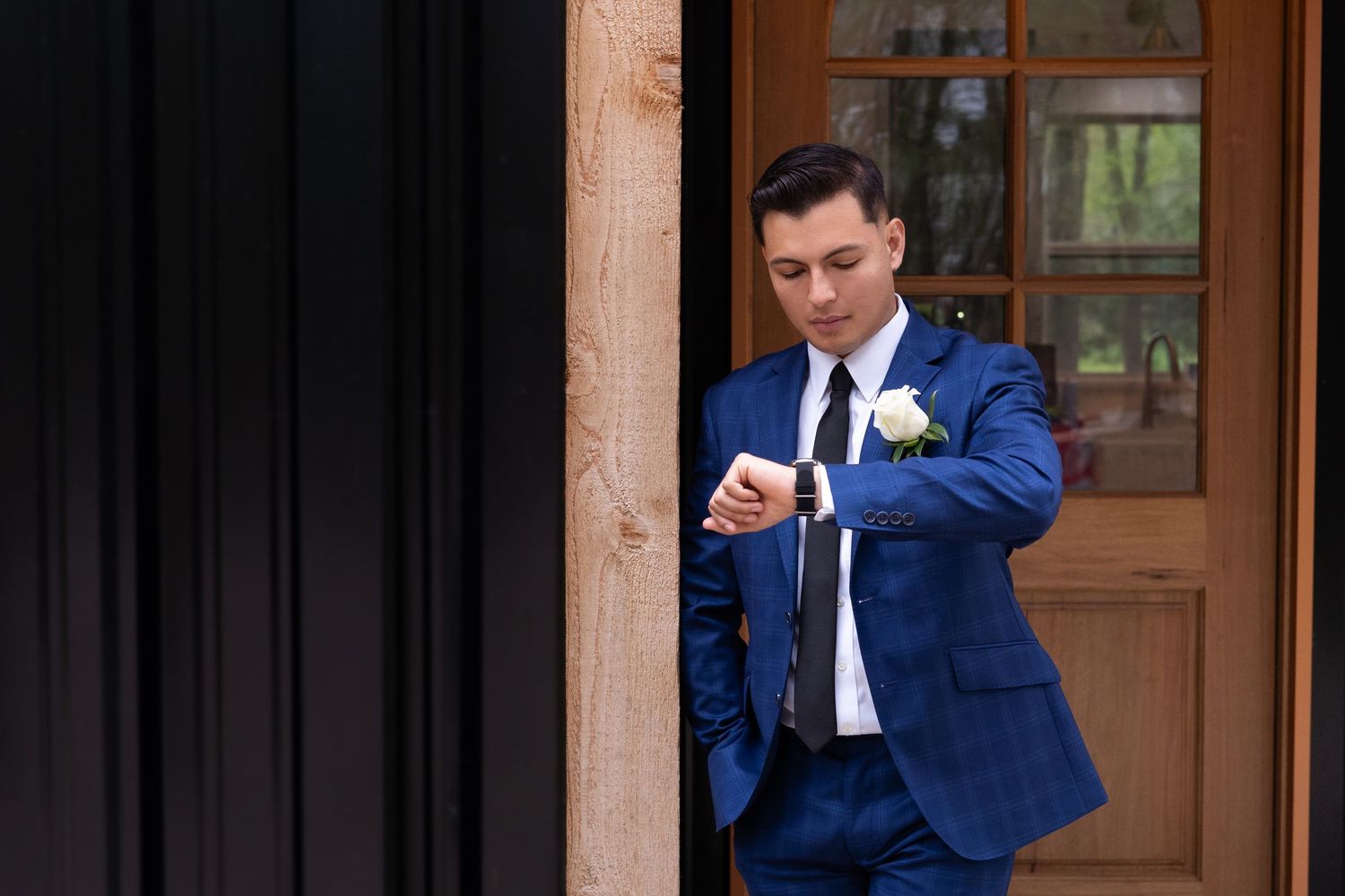 Groom in navy blue suit checks his watch while standing by wooden doors at wedding venue.