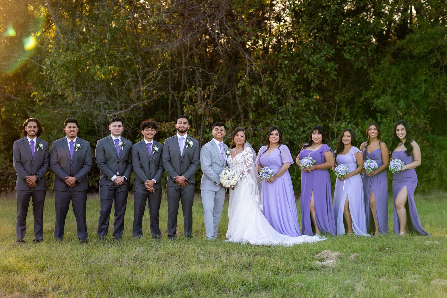 Wedding party poses outdoors with bridesmaids in purple dresses and groomsmen in grey suits.