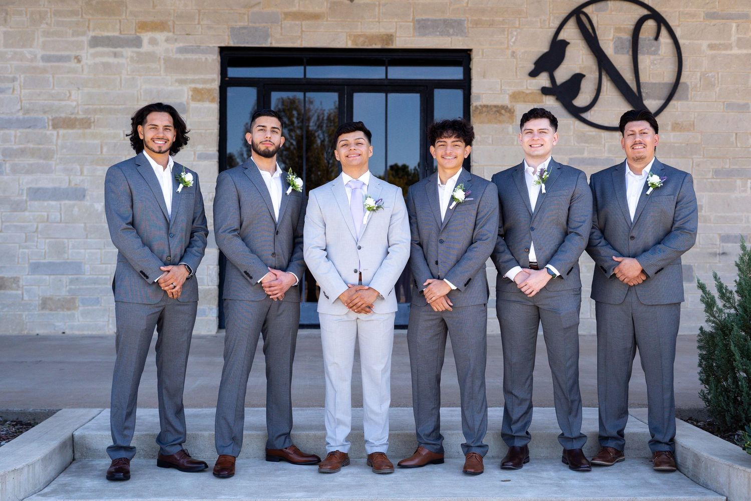 Groomsmen in gray suits and one in white stand together on stone steps outside a building.