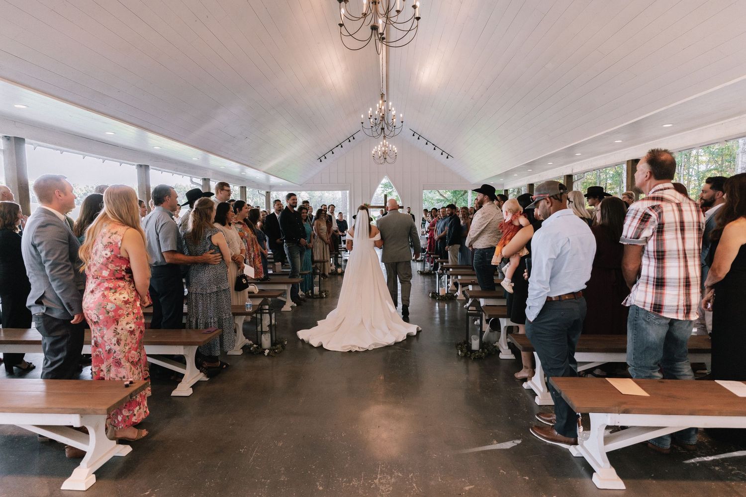 Wedding ceremony venue with elegant chandeliers and white decor features rows of guests seated in a bright space.