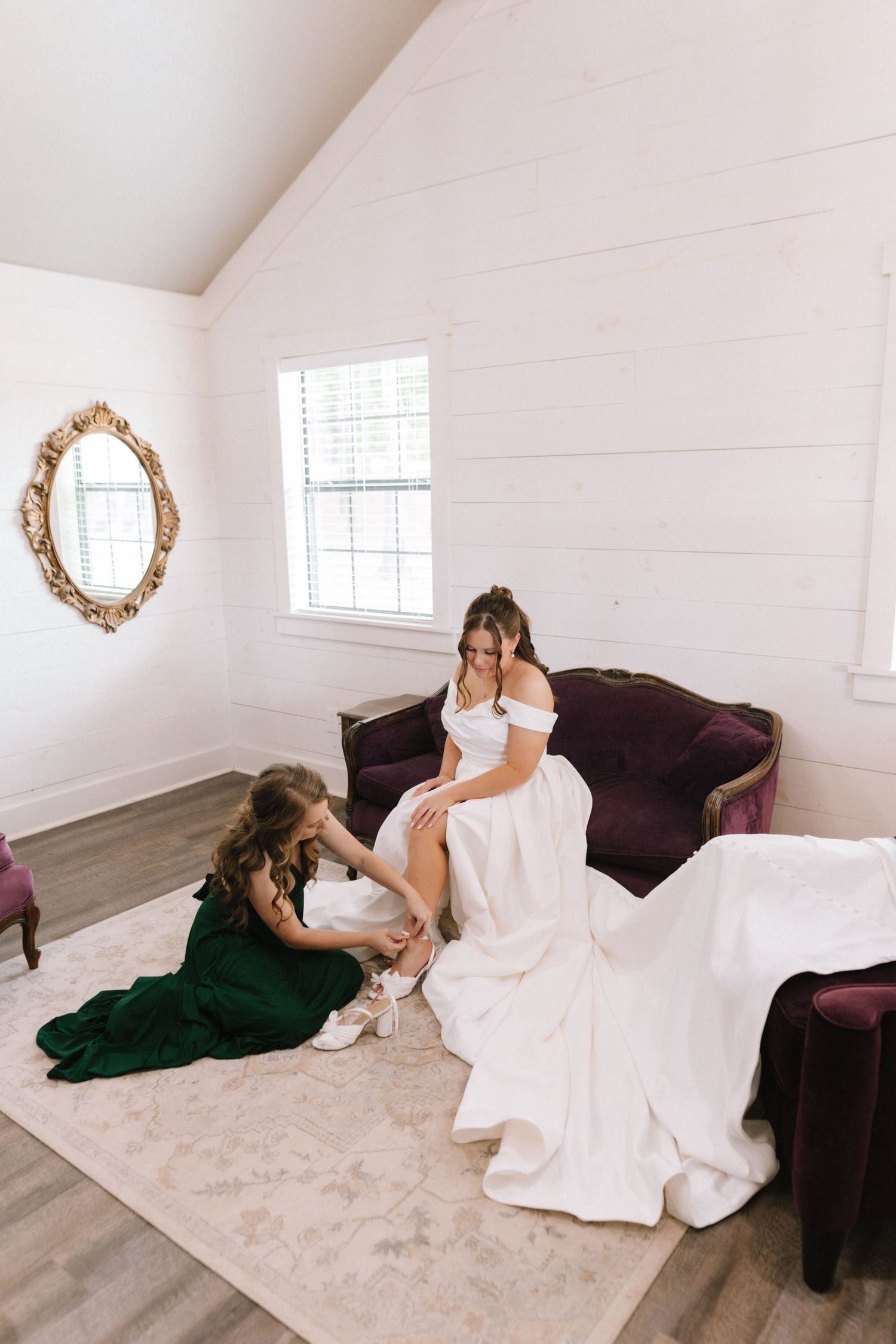 Two people in formal attire share a tender moment on a purple couch in a bright white room with shiplap walls.