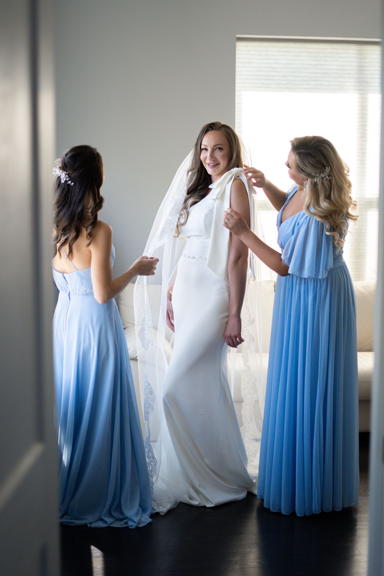 Two bridesmaids in flowing blue dresses help adjust a bride's veil in an elegant getting-ready moment.