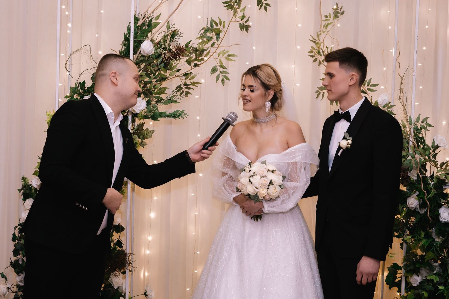 A wedding ceremony takes place in front of a floral arch with twinkle lights as the officiant speaks into a microphone.