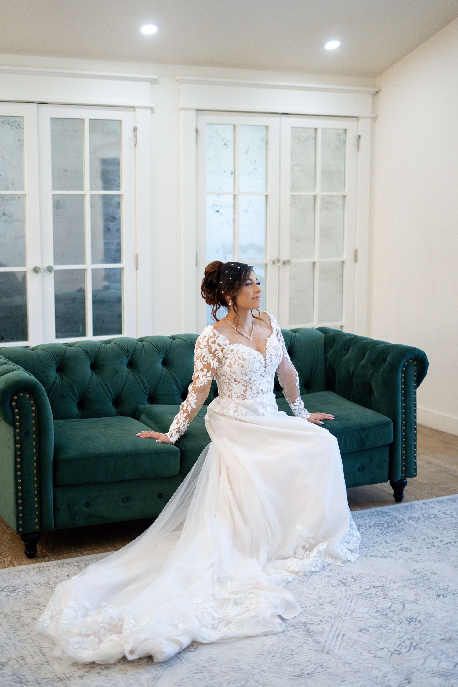 Elegant bride in lace wedding dress poses on emerald green tufted couch in bright indoor setting.