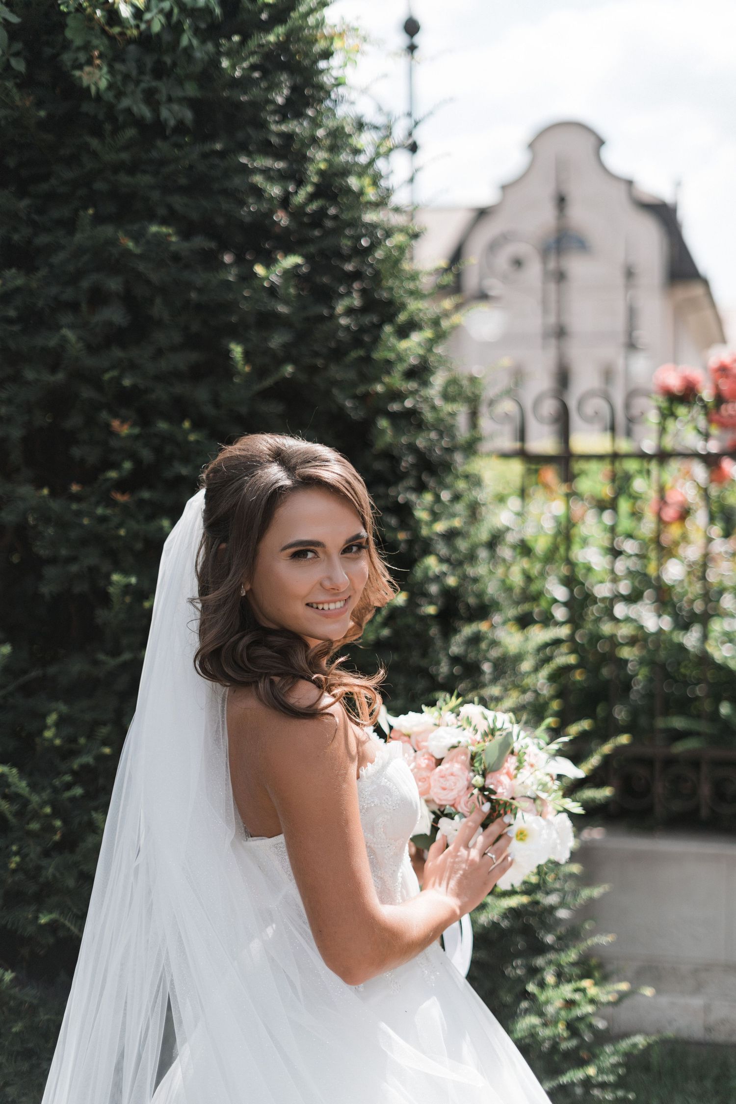 A bride in a white wedding dress poses with a bouquet in a garden setting with architectural details in the background.