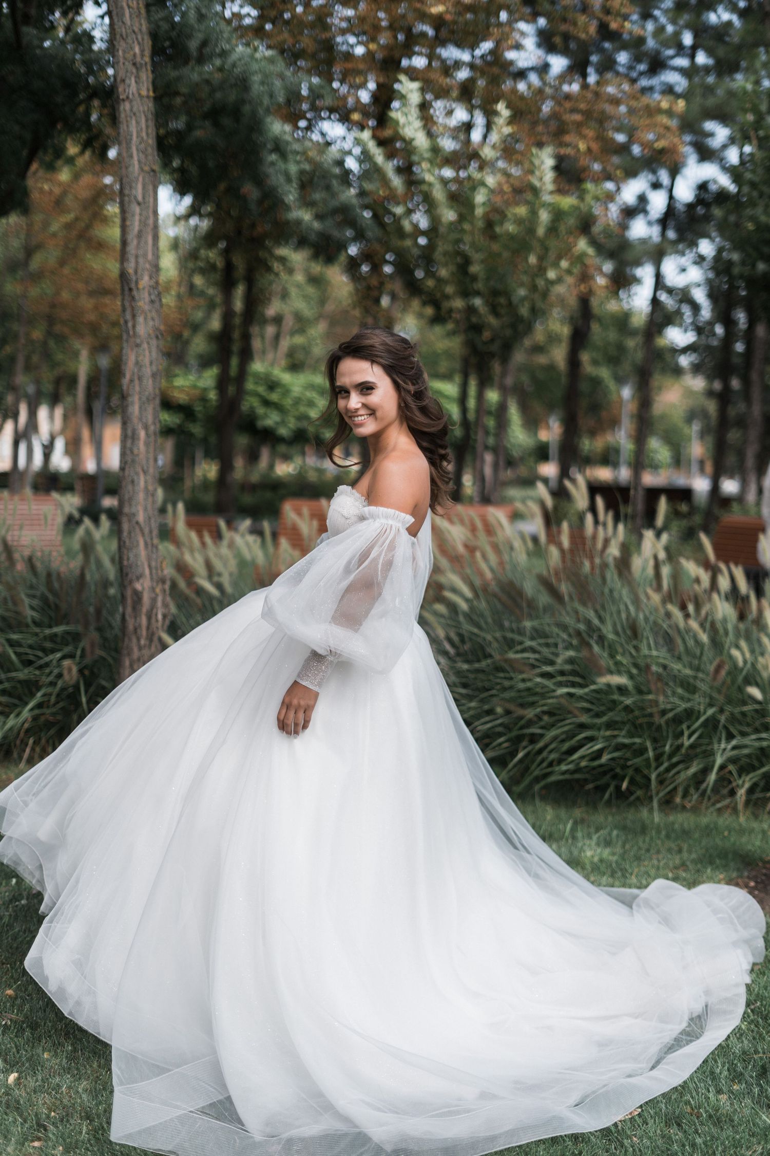 A bride in a flowing white wedding gown twirls gracefully among tall pine trees in an outdoor setting.