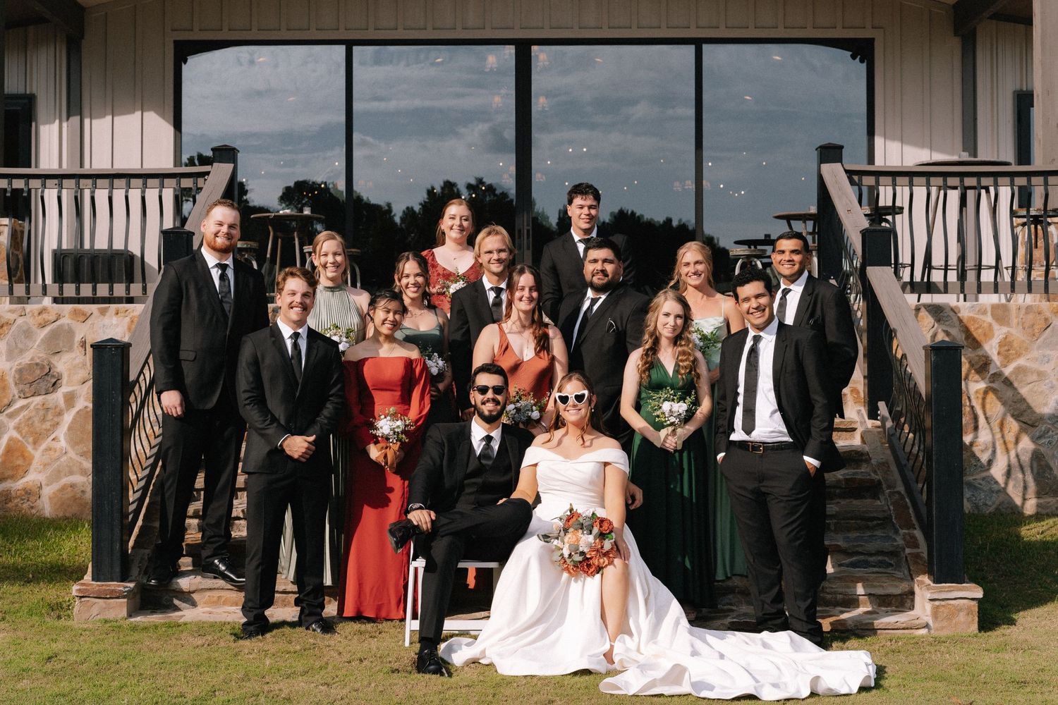 Large wedding party group photo taken outside with stone and white architectural elements.