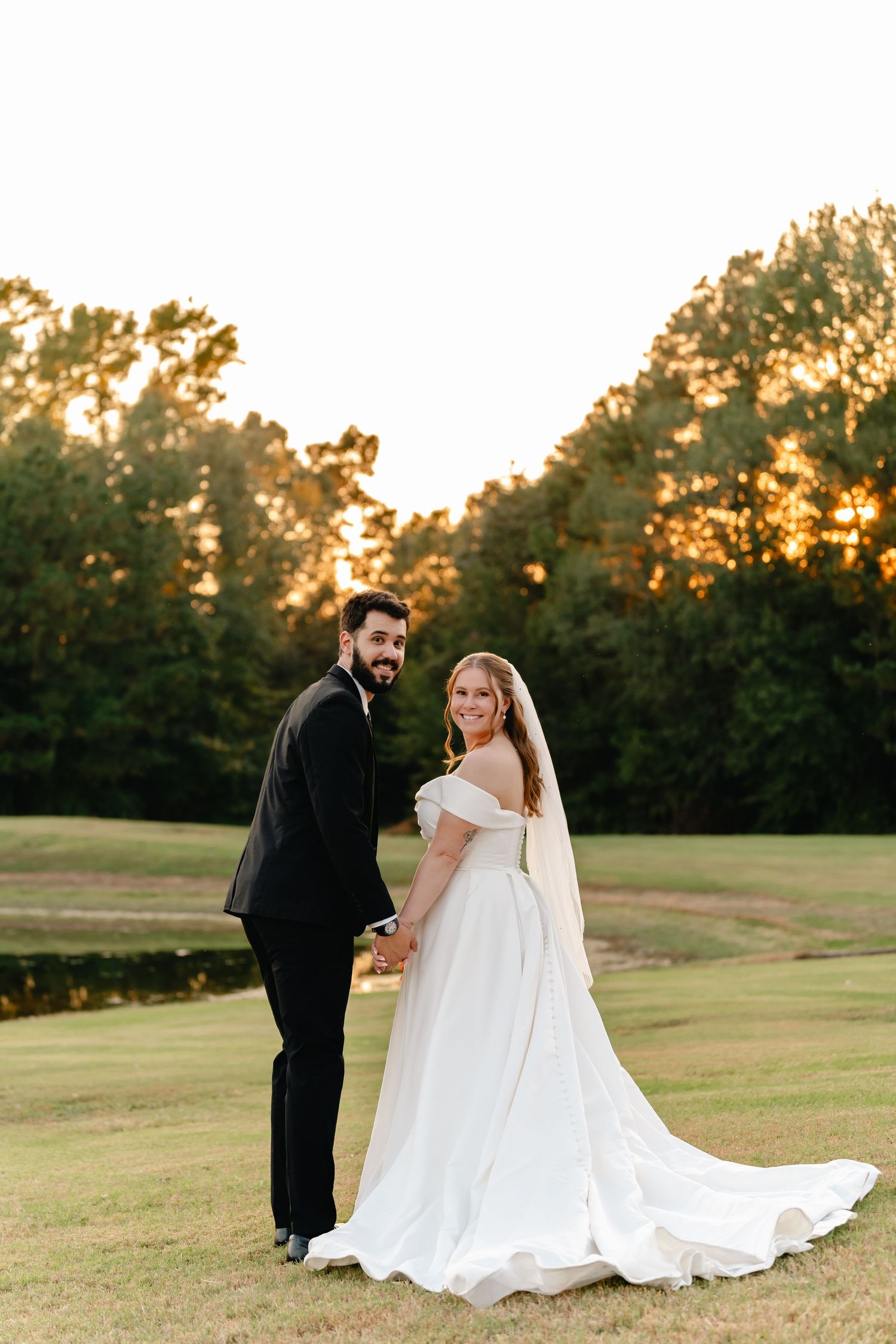 A sunset wedding portrait of a couple standing on a grassy field with golden light filtering through trees.
