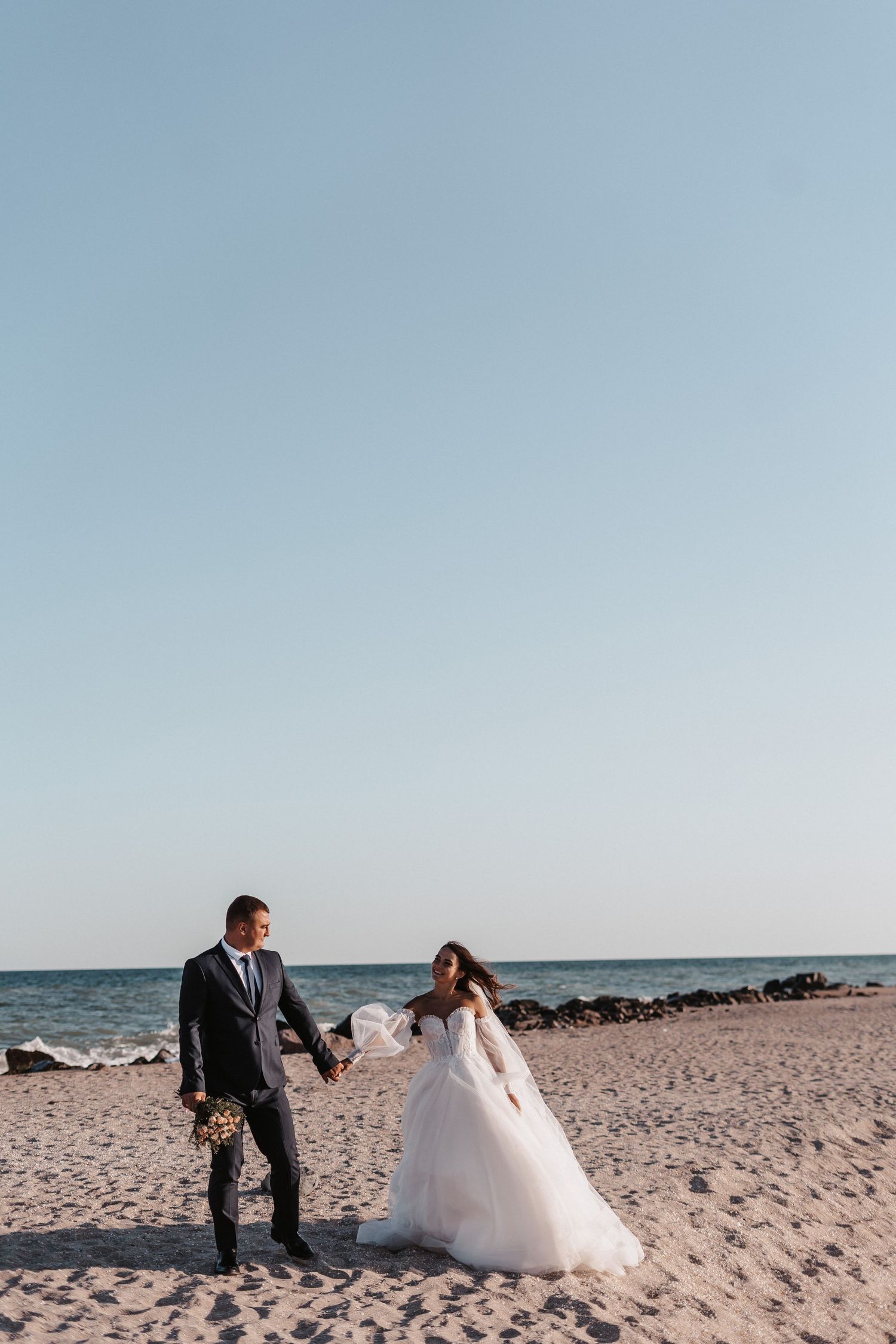 Romantic beach wedding photoshoot with couple walking along sandy shoreline at sunset.