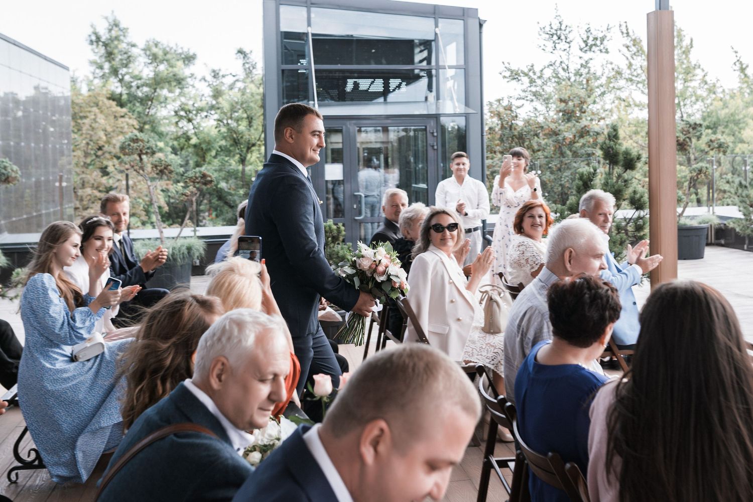 A group of people gather at an outdoor event space with glass walls and natural lighting.