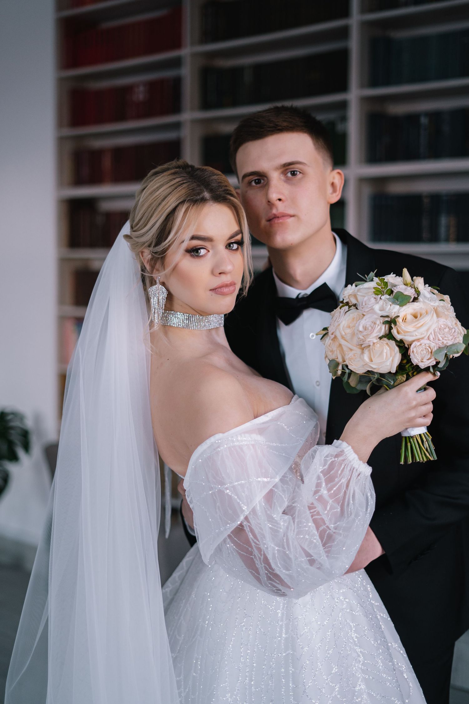 Romantic wedding couple poses together in formal attire with pink rose bouquet and flowing white veil.