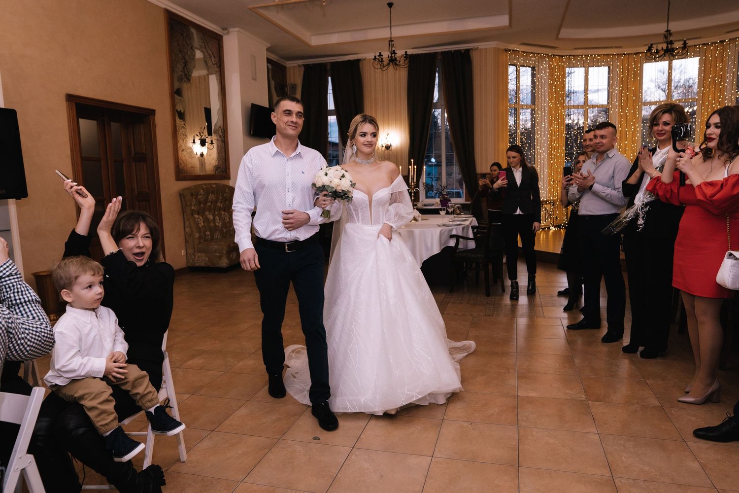 A wedding party stands in line in an elegant ballroom hallway with guests watching from the sides.