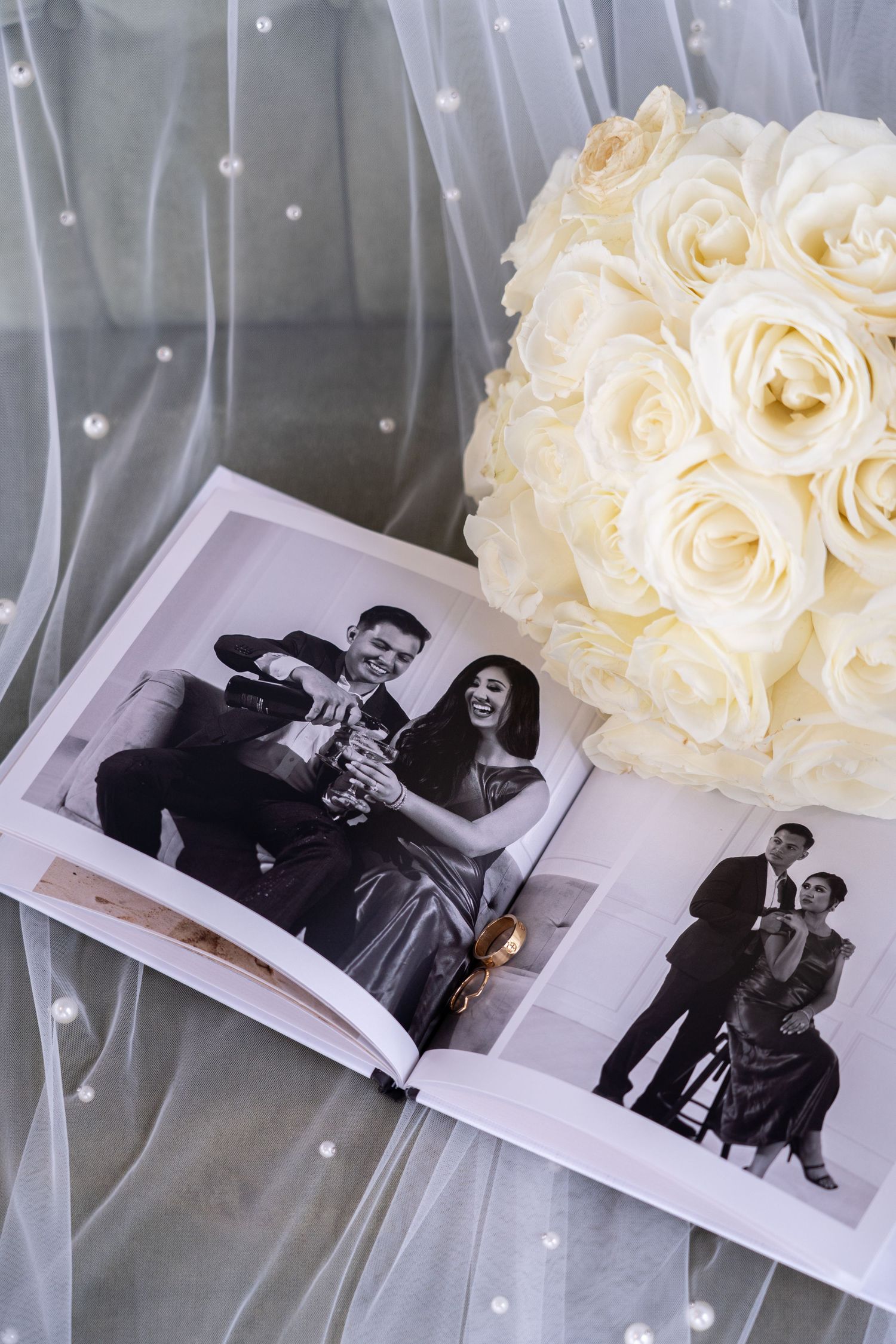 A wedding photo album lies open next to a bouquet of cream-colored roses on white tulle fabric.