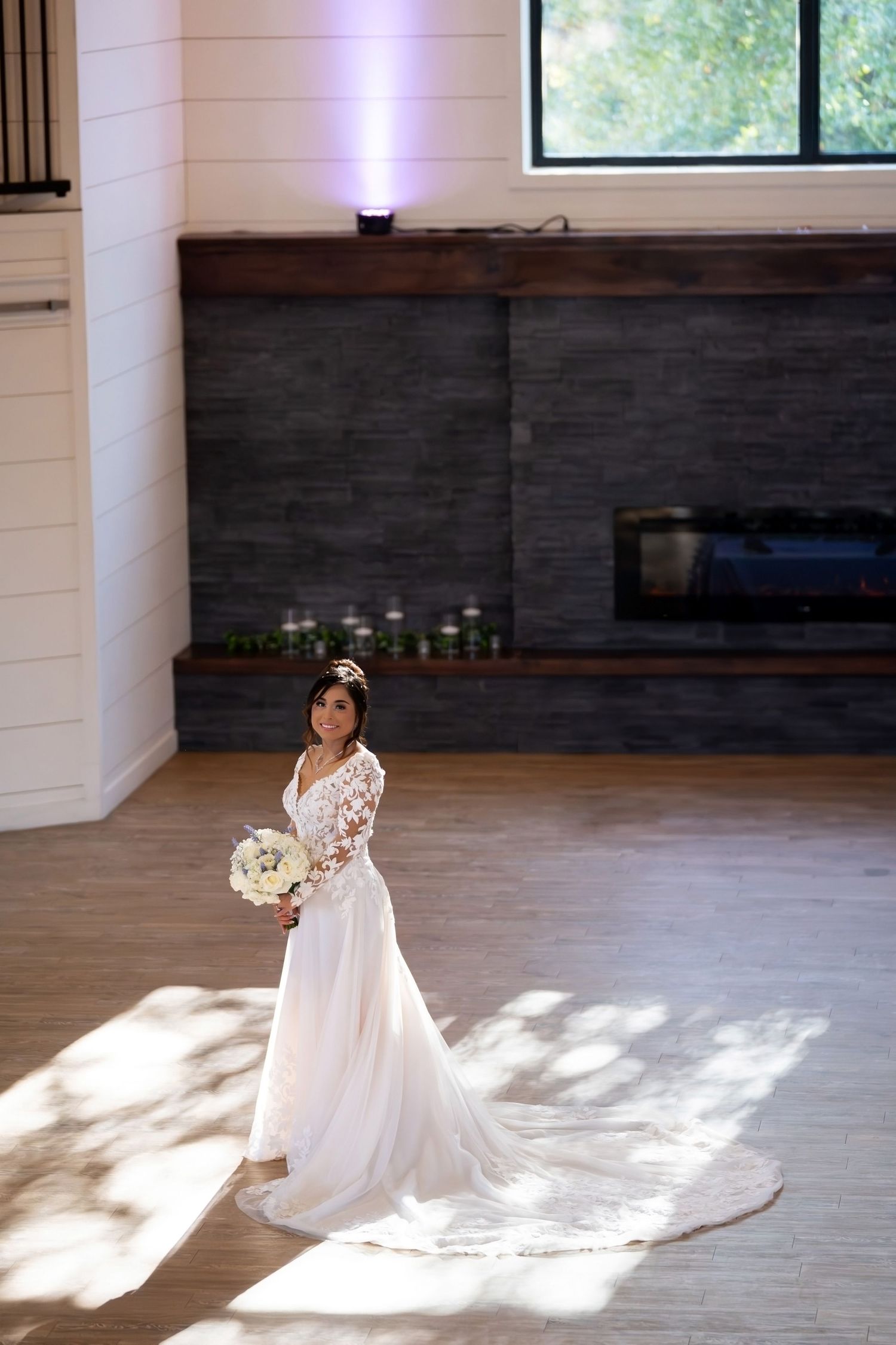 Elegant bride in flowing white dress poses in sunlit room with wooden floors and dark accent walls.