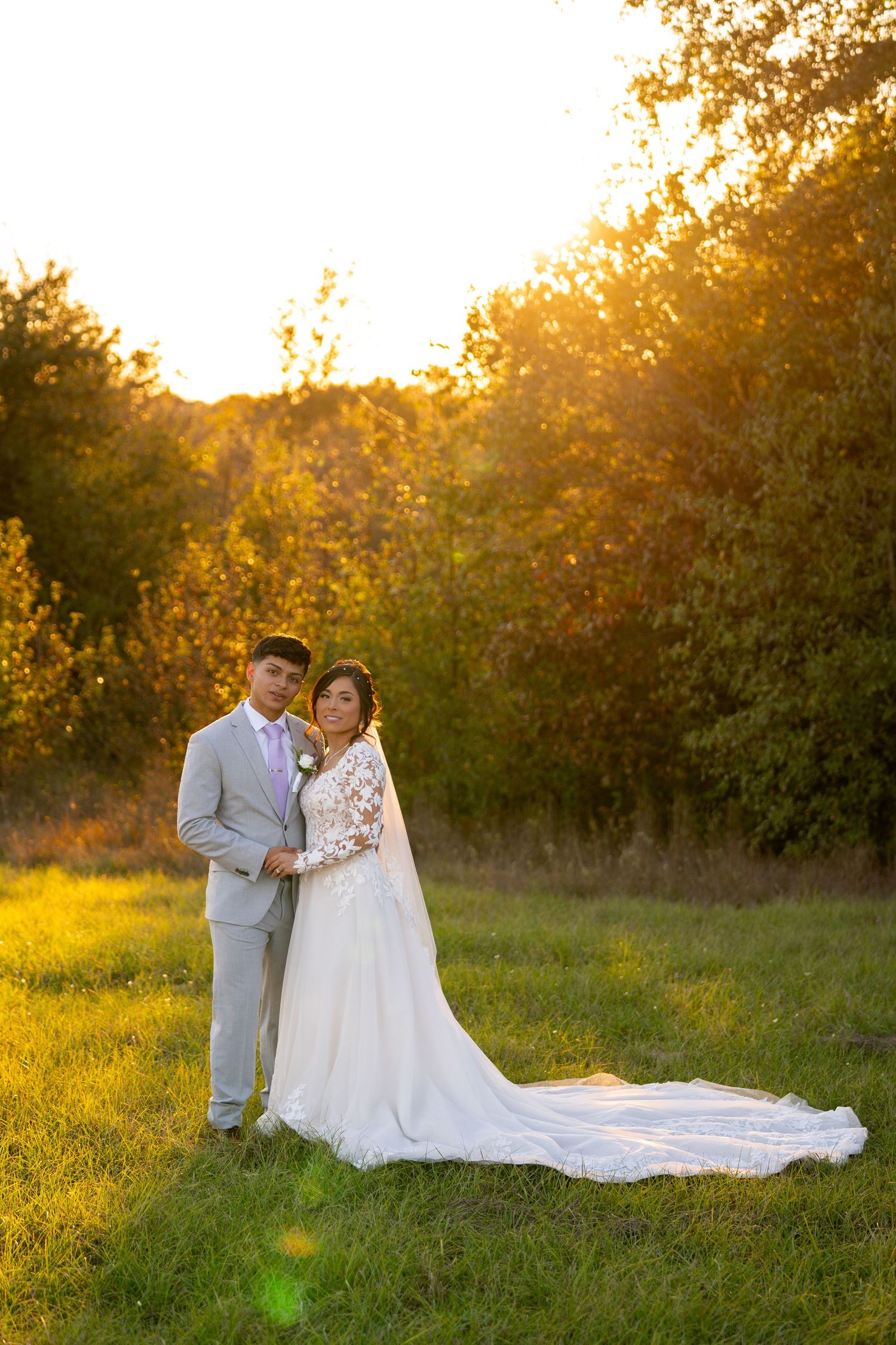 Couple in wedding attire poses in a sunlit meadow during golden hour with a flowing white dress train.