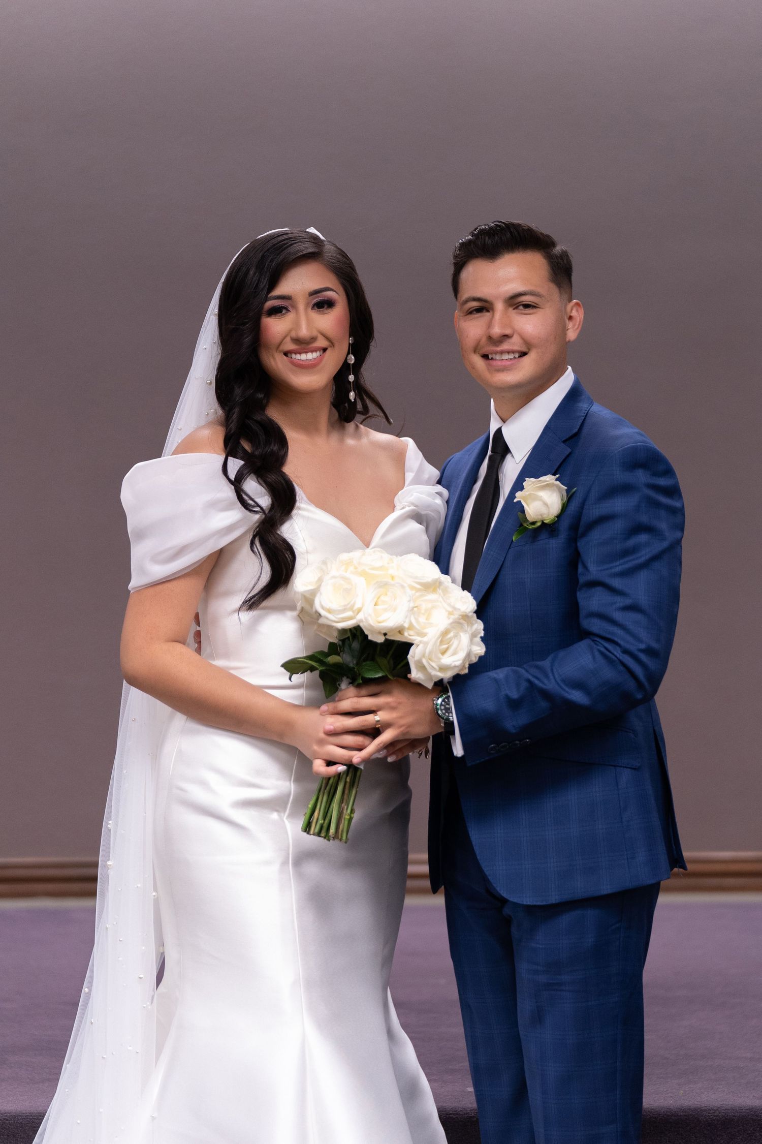 A bride in a white dress and groom in a navy suit pose together for wedding photos holding a white rose bouquet.