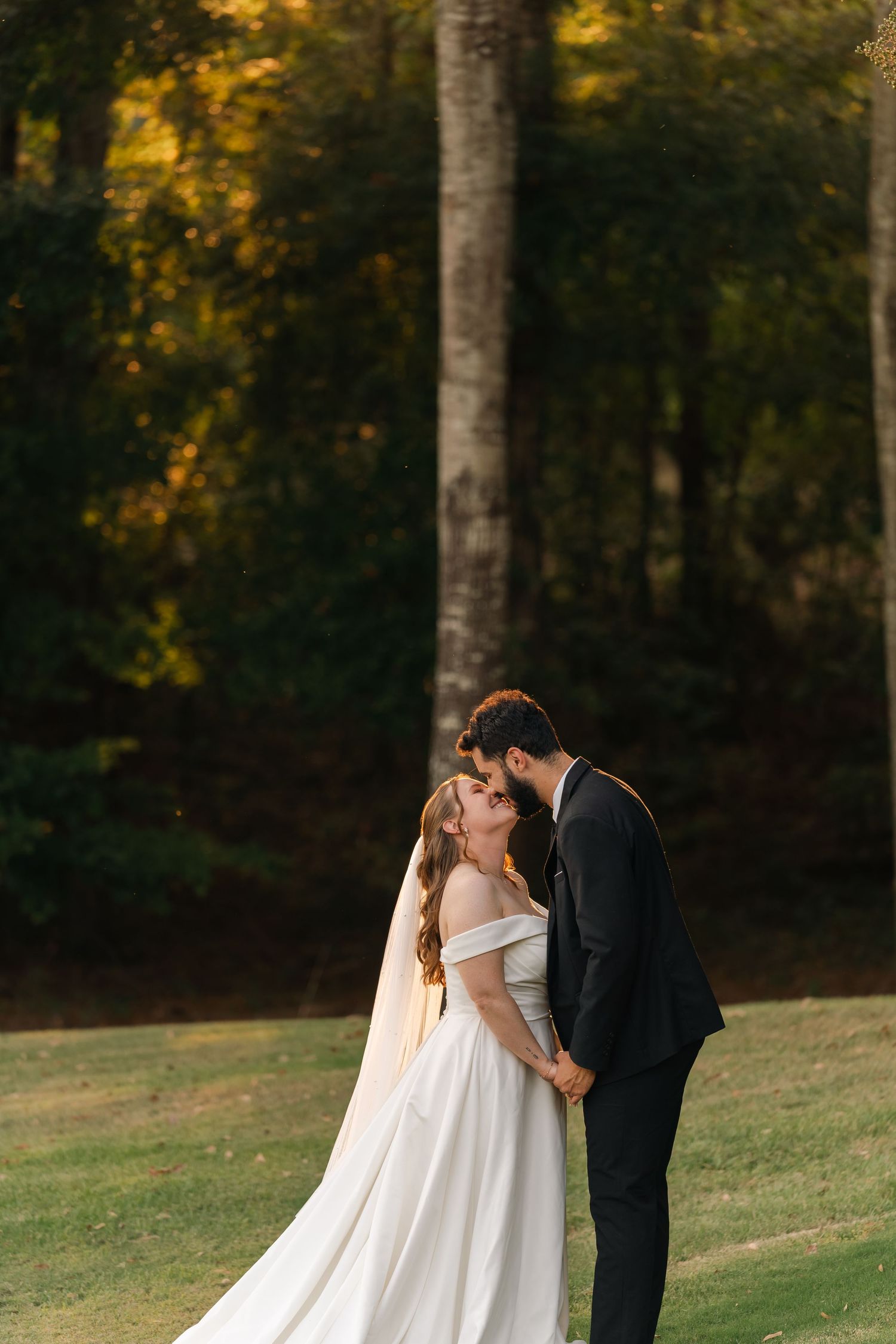 A bride in an off-shoulder white gown and groom in black suit share intimate moments during their outdoor wedding.