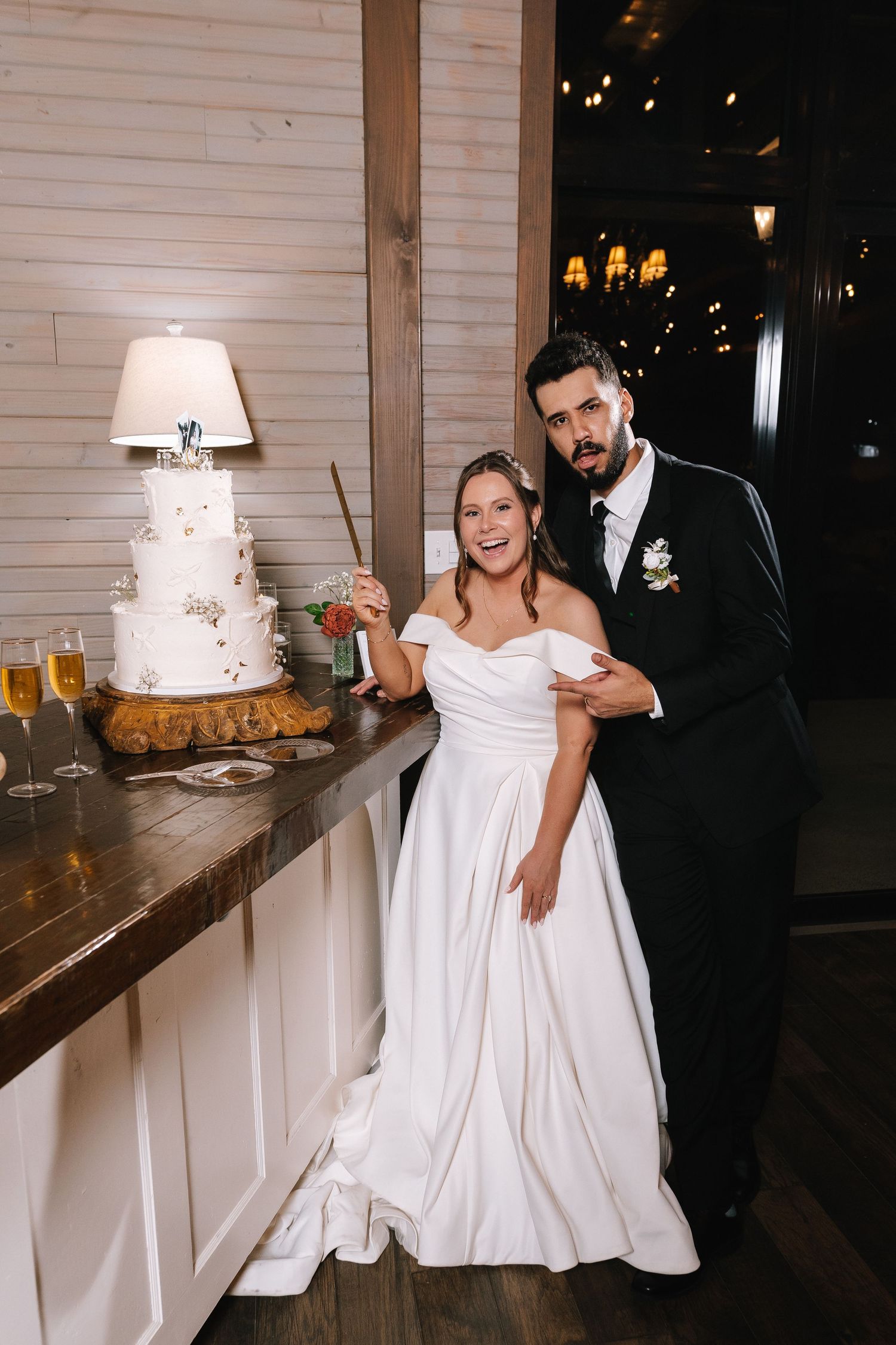 Wedding couple cuts their elegant white tiered cake at rustic wooden reception venue.