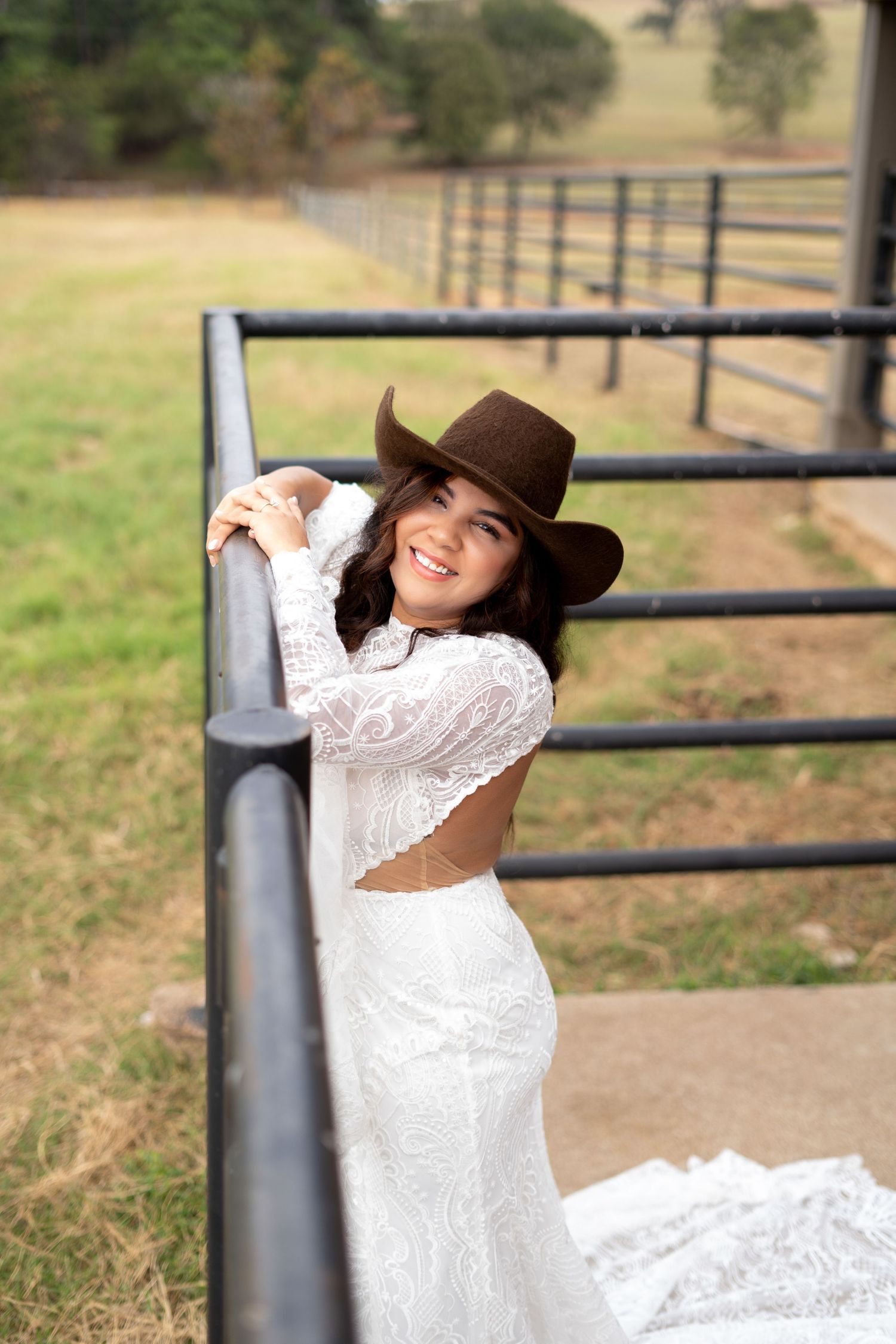 Bride in lace dress and brown hat poses playfully against rustic fence.