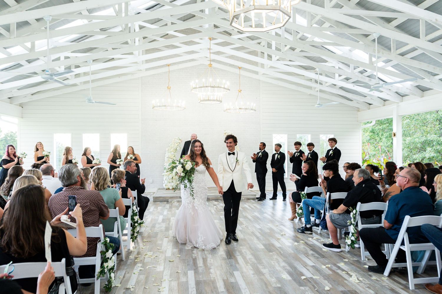 Wedding guests gather under a bright white tent with elegant lighting during an outdoor ceremony.