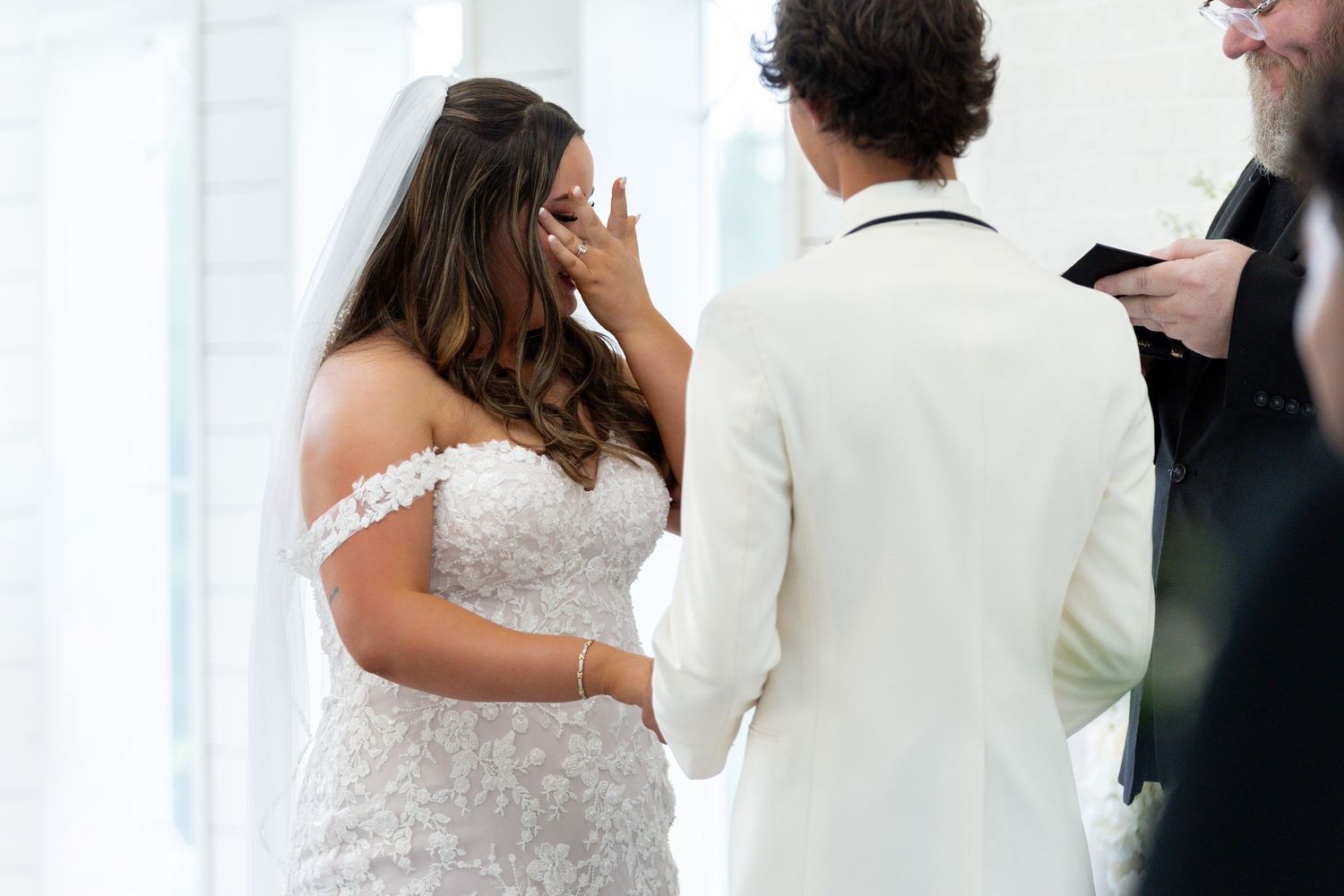 Emotional moment during wedding ceremony as bride and groom exchange vows in front of white curtains.