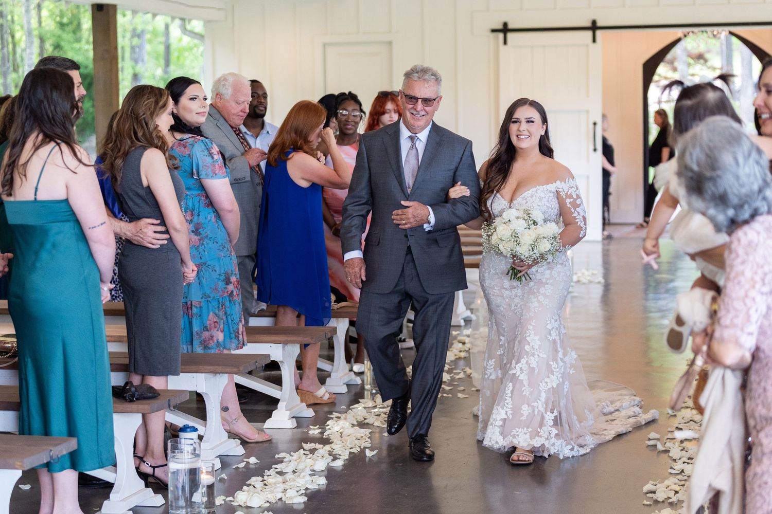 Wedding party members walk down the aisle decorated with white flower petals during an indoor ceremony.