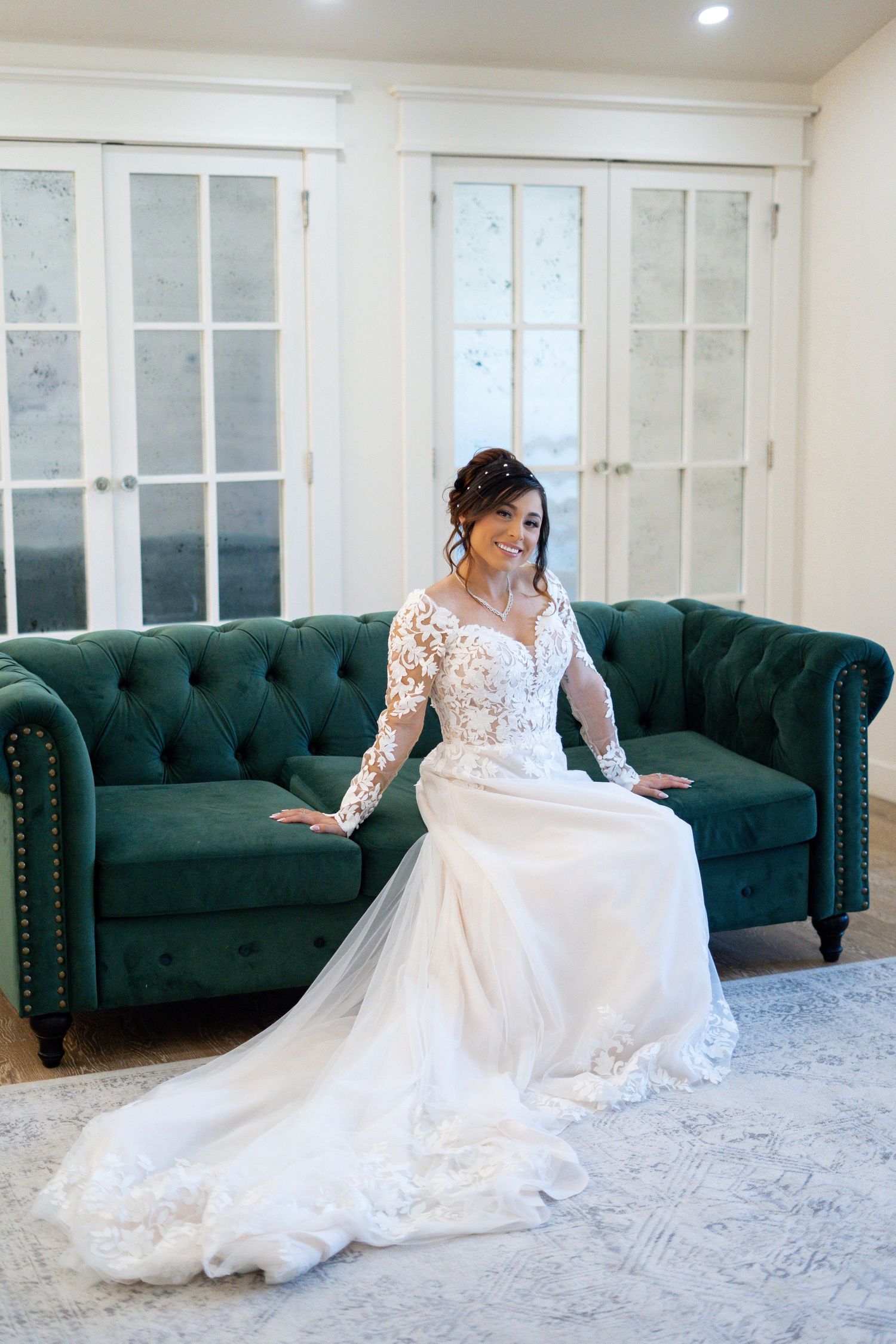 Elegant bride in long sleeve lace wedding gown sits gracefully on emerald green tufted sofa in bright room.