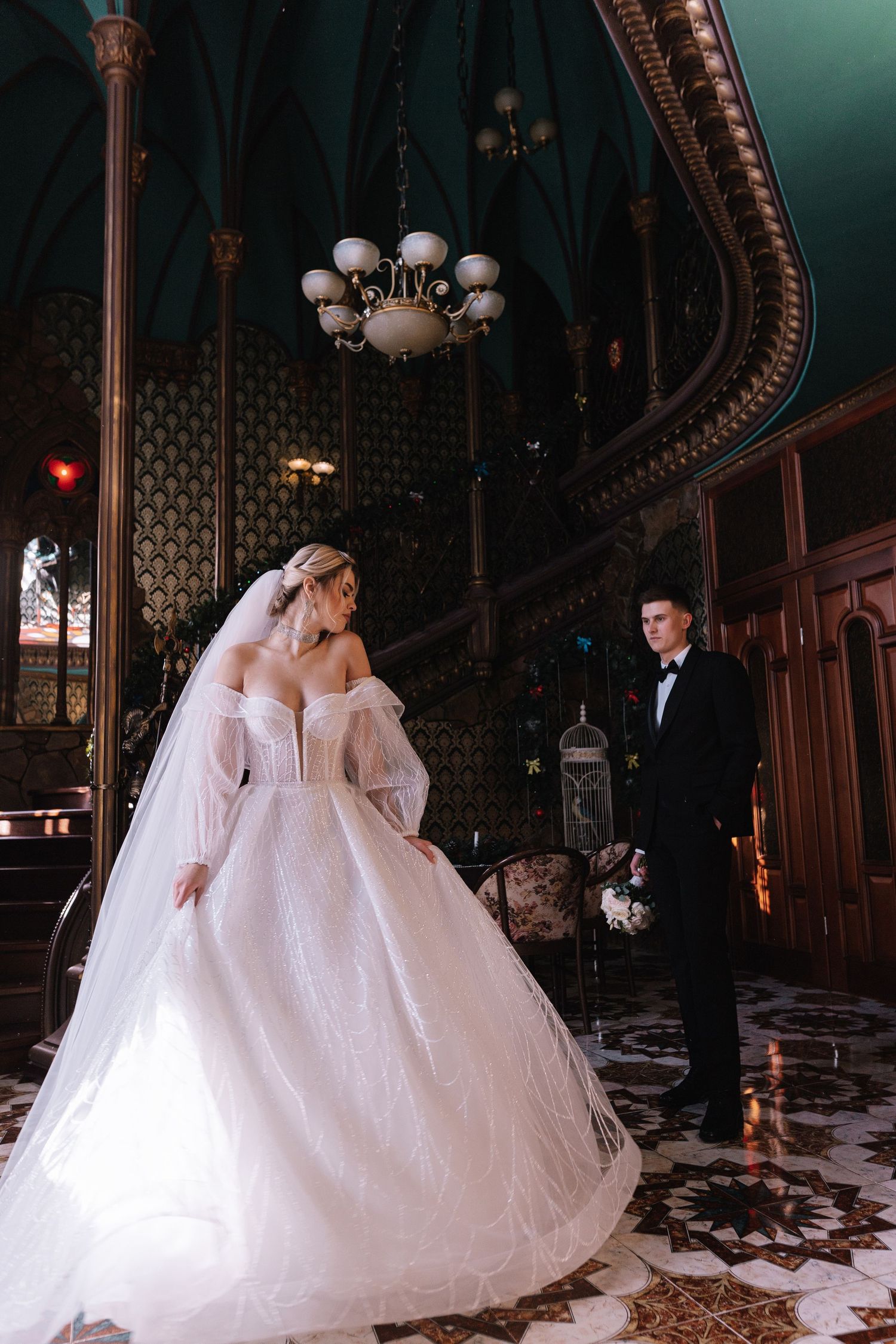 A couple in wedding attire shares a romantic moment in a grand vintage-style ballroom with ornate architectural details.