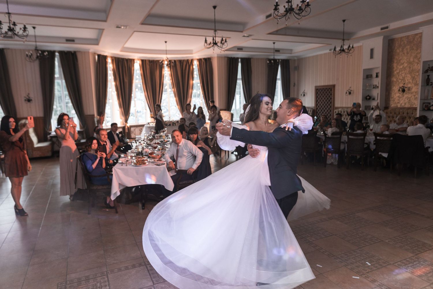 Wedding guests dance and celebrate in an elegant ballroom with white decor and chandeliers.