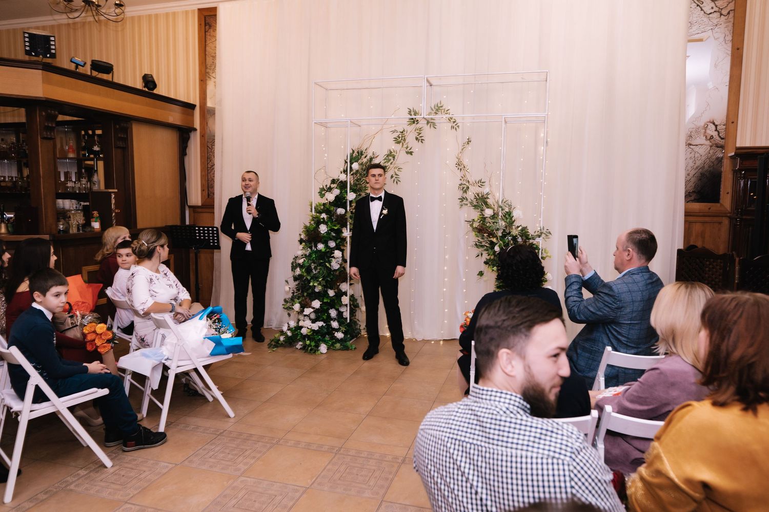Wedding guests seated in rows of white chairs at an indoor ceremony venue with exposed brick walls.