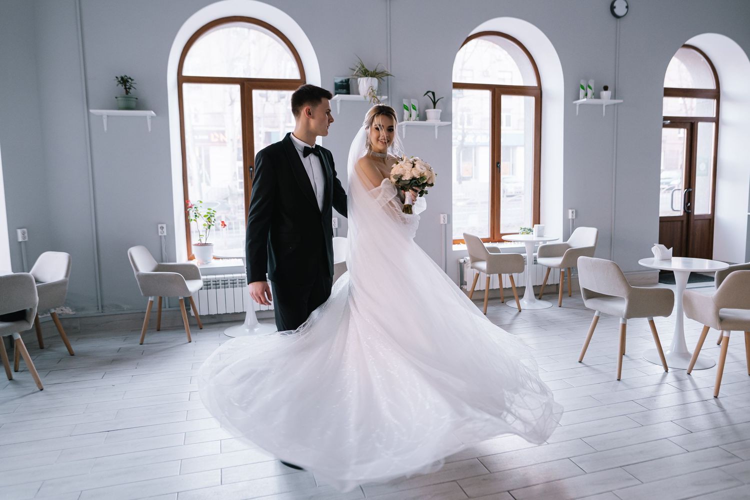 A couple in formal wedding attire dancing in an elegant white room with arched windows and modern decor.