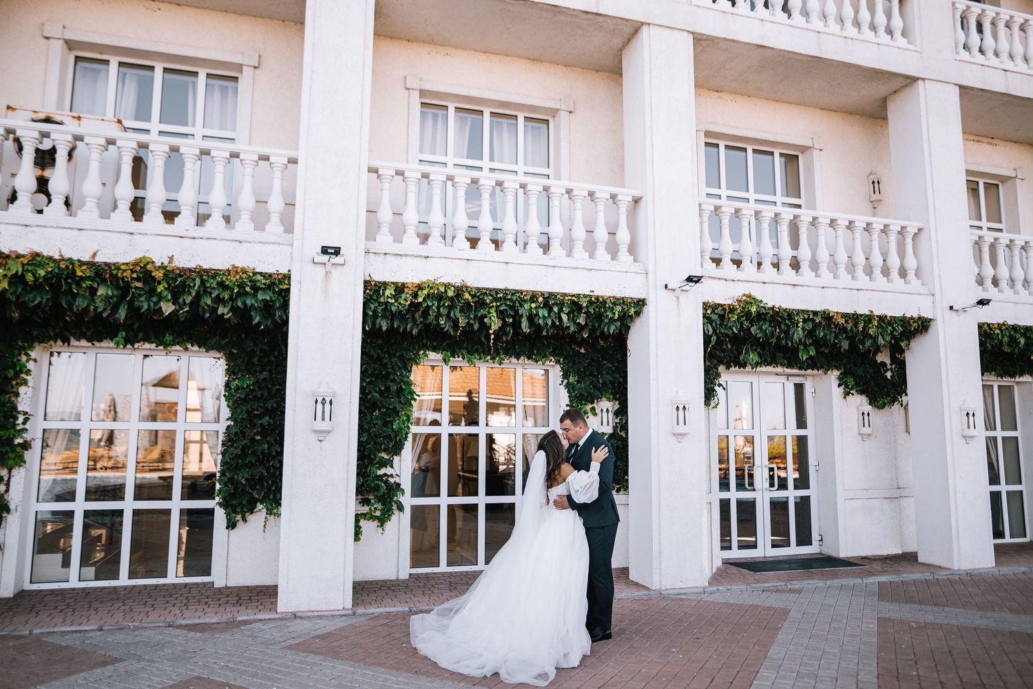 White columns and balustrades line the exterior of an elegant wedding venue with glass windows and greenery accents.