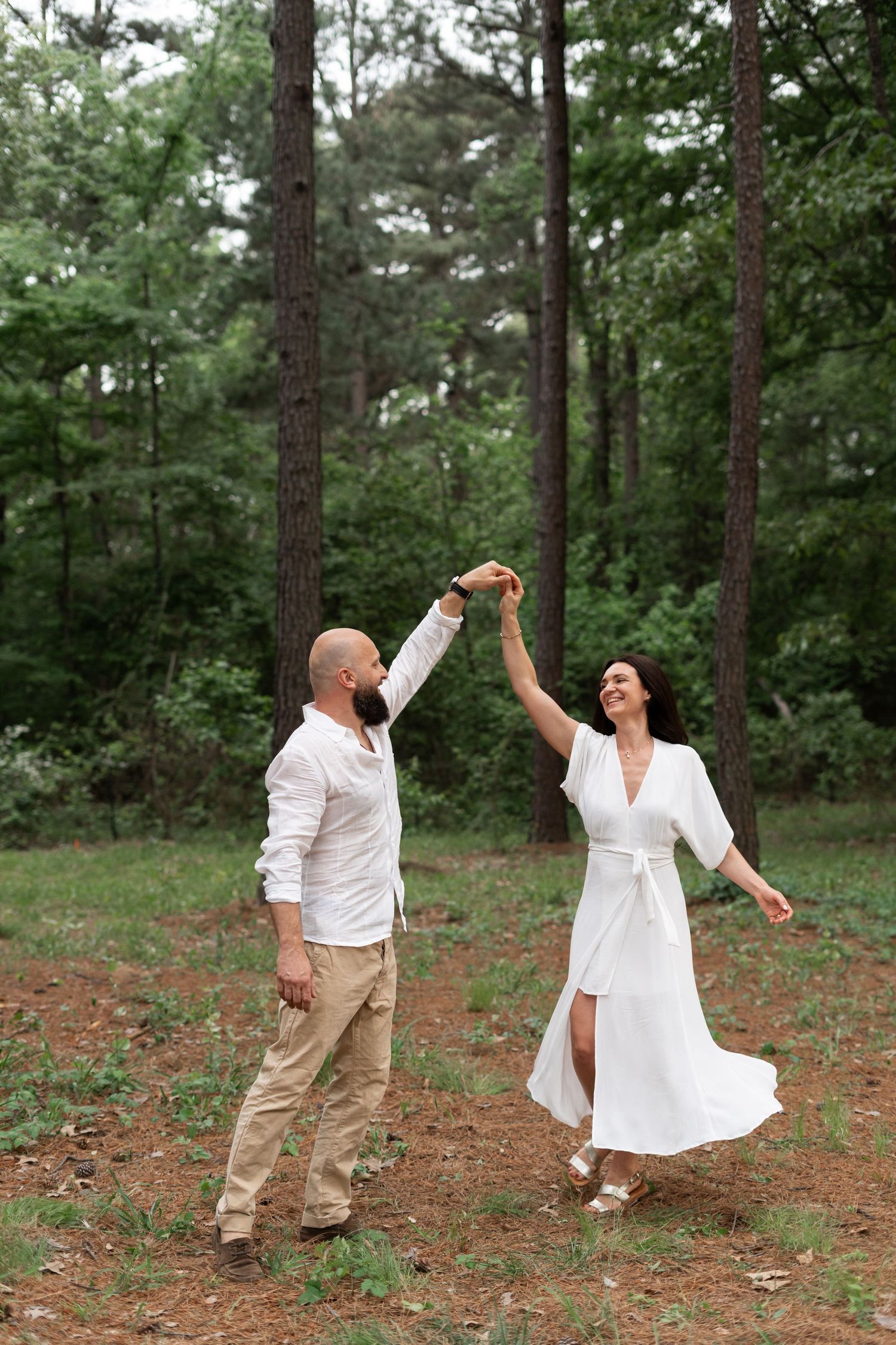 A couple in white outfits dances joyfully in a pine forest setting.