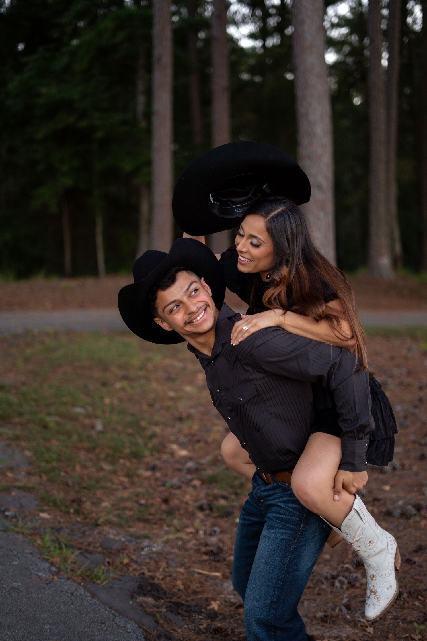 A playful piggyback ride moment between a couple in western attire near pine trees.