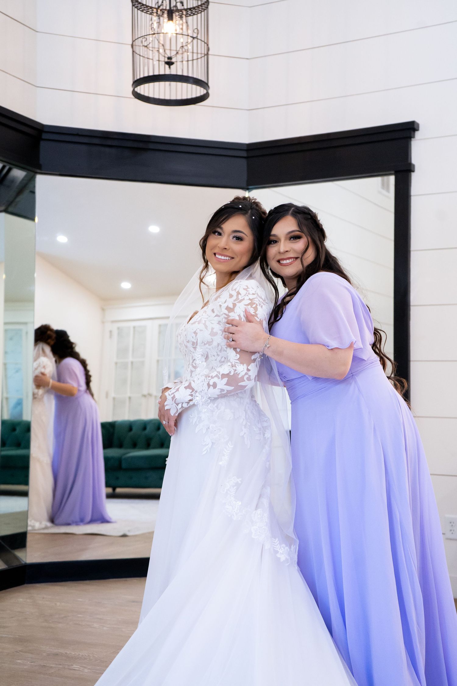 A bride in a white lace dress poses with someone wearing a lavender gown in an elegant indoor setting.
