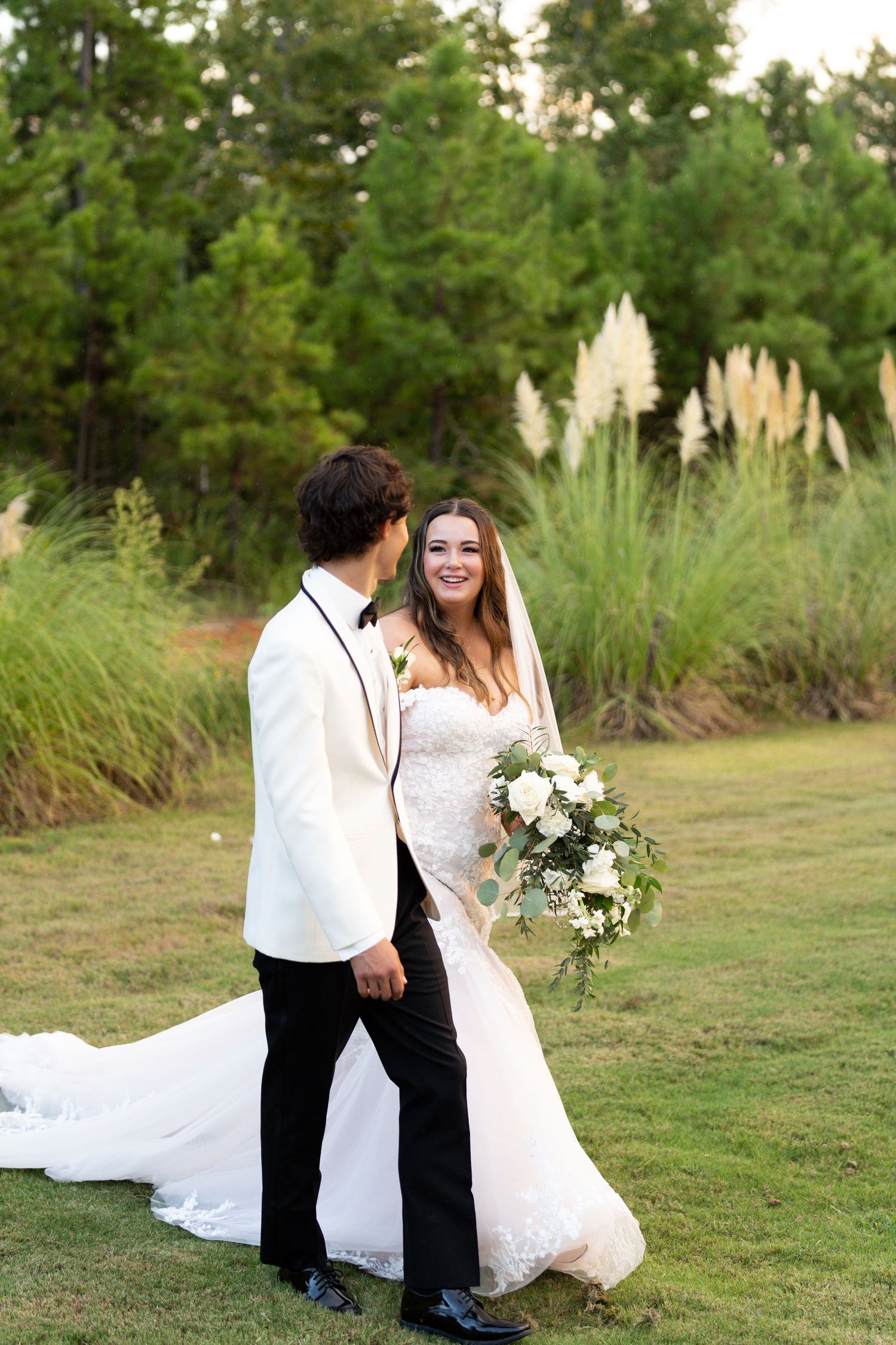 A couple in wedding attire stands together on a lawn with pine trees and pampas grass in the background.
