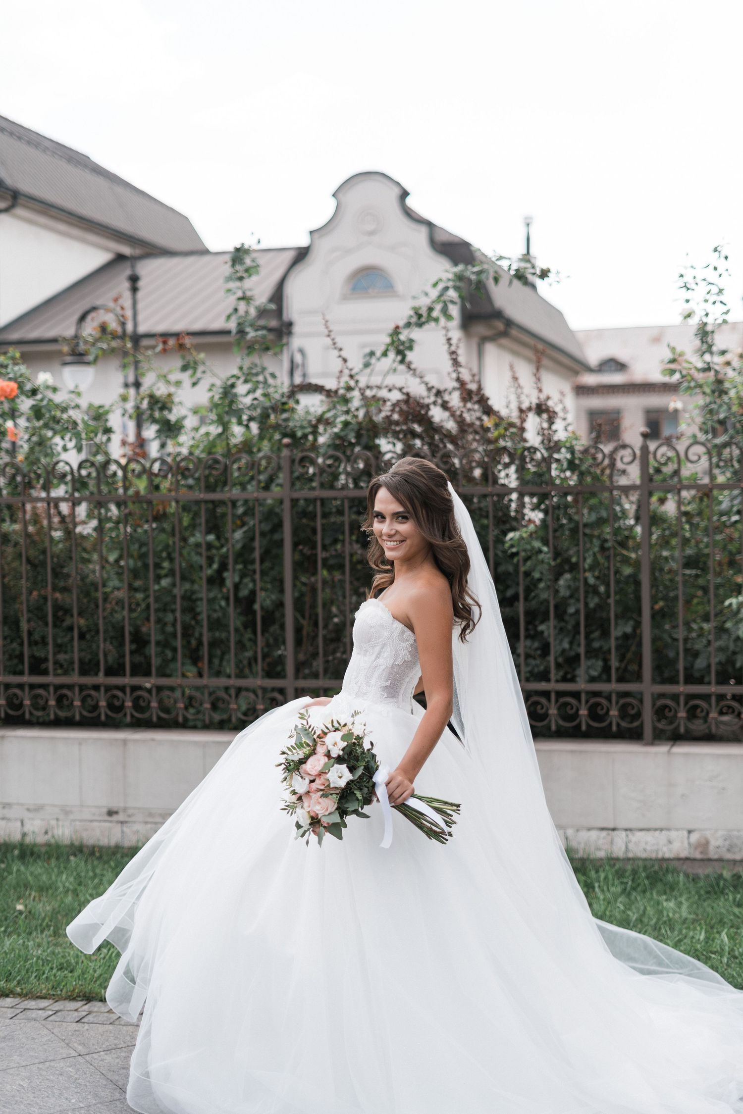 A bride in a flowing white wedding dress twirls gracefully on a manicured lawn beside a wrought iron fence.