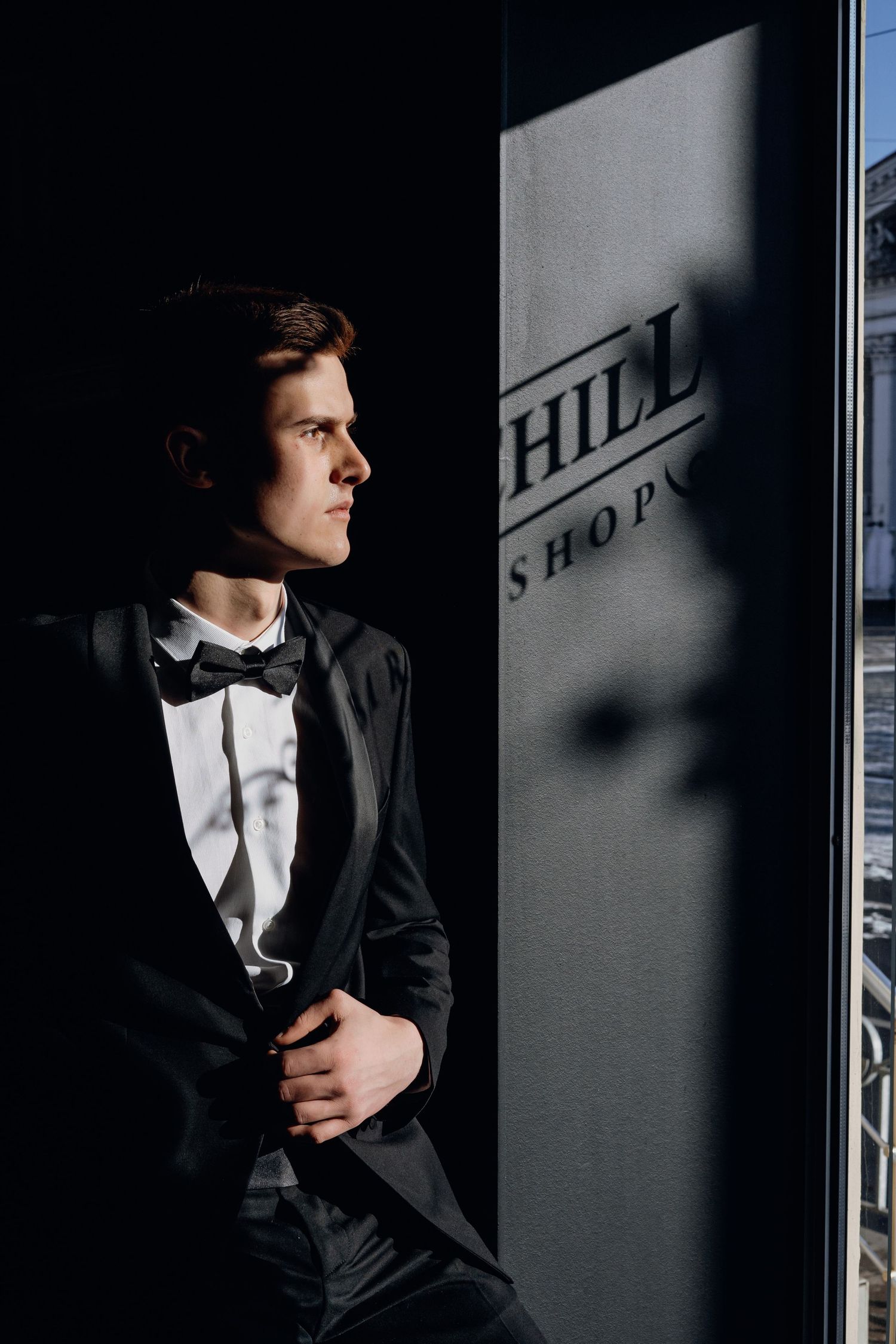 A person in formal black tuxedo and bow tie stands in dramatic lighting against a dark wall with shadows.