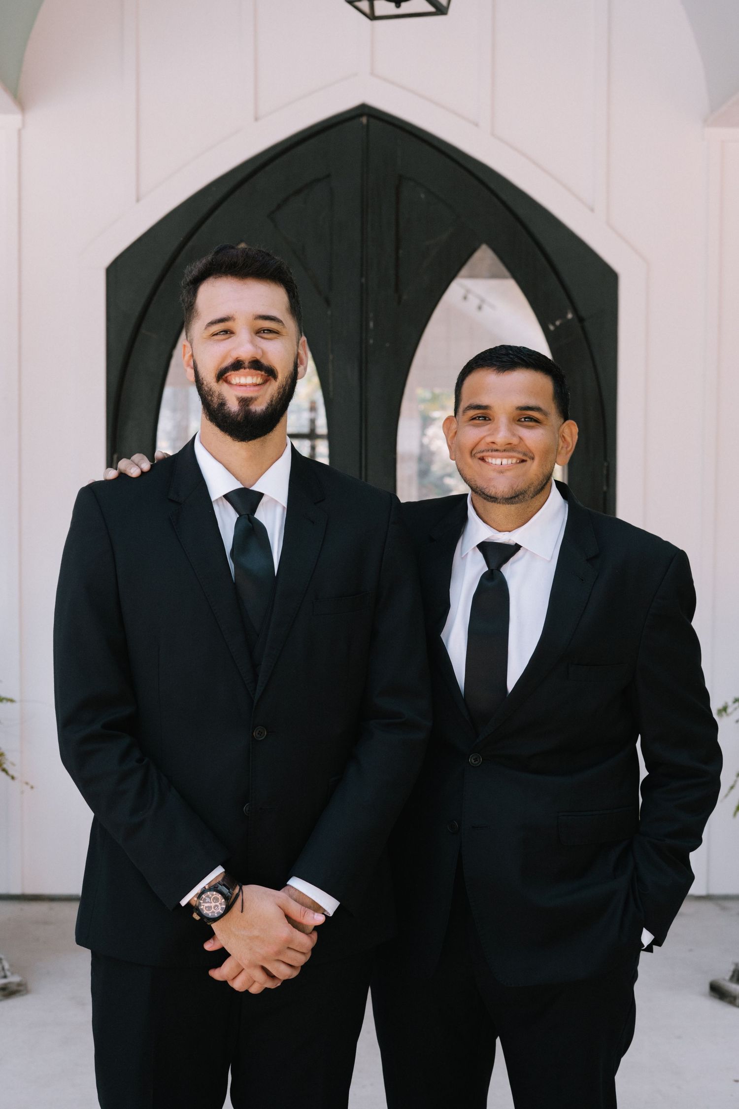 Groomsmen in black suits and ties posing together against white chapel backdrop.