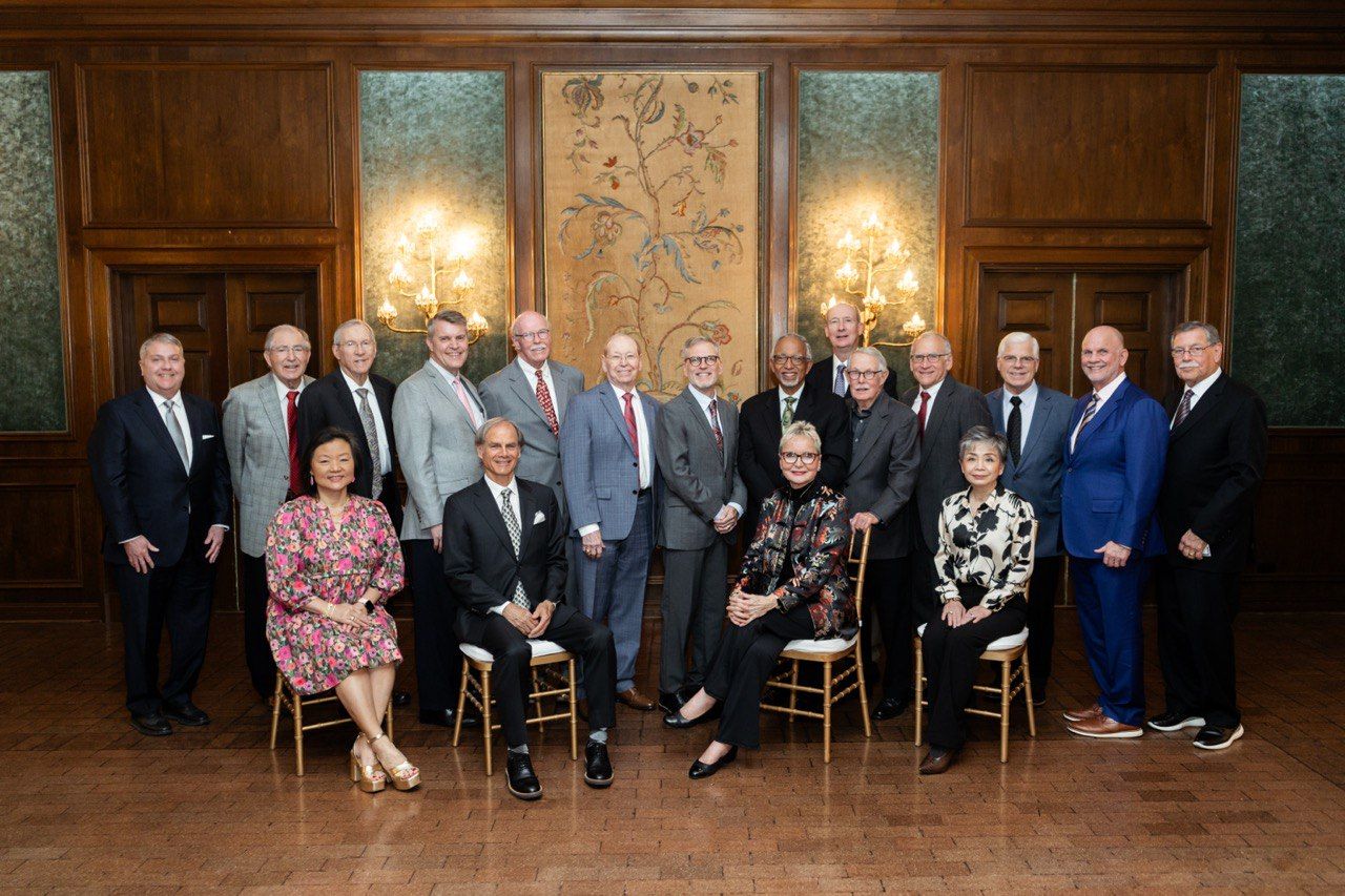 Formal group portrait of elegantly dressed guests seated in a grand wood-paneled room with ornate decor.