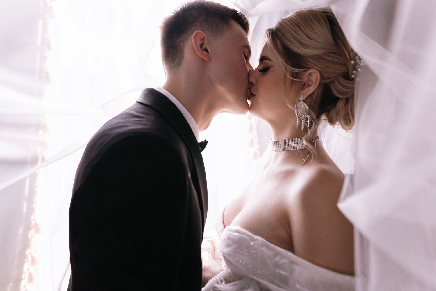 A romantic wedding moment captured through sheer white curtains as a couple shares a tender kiss.