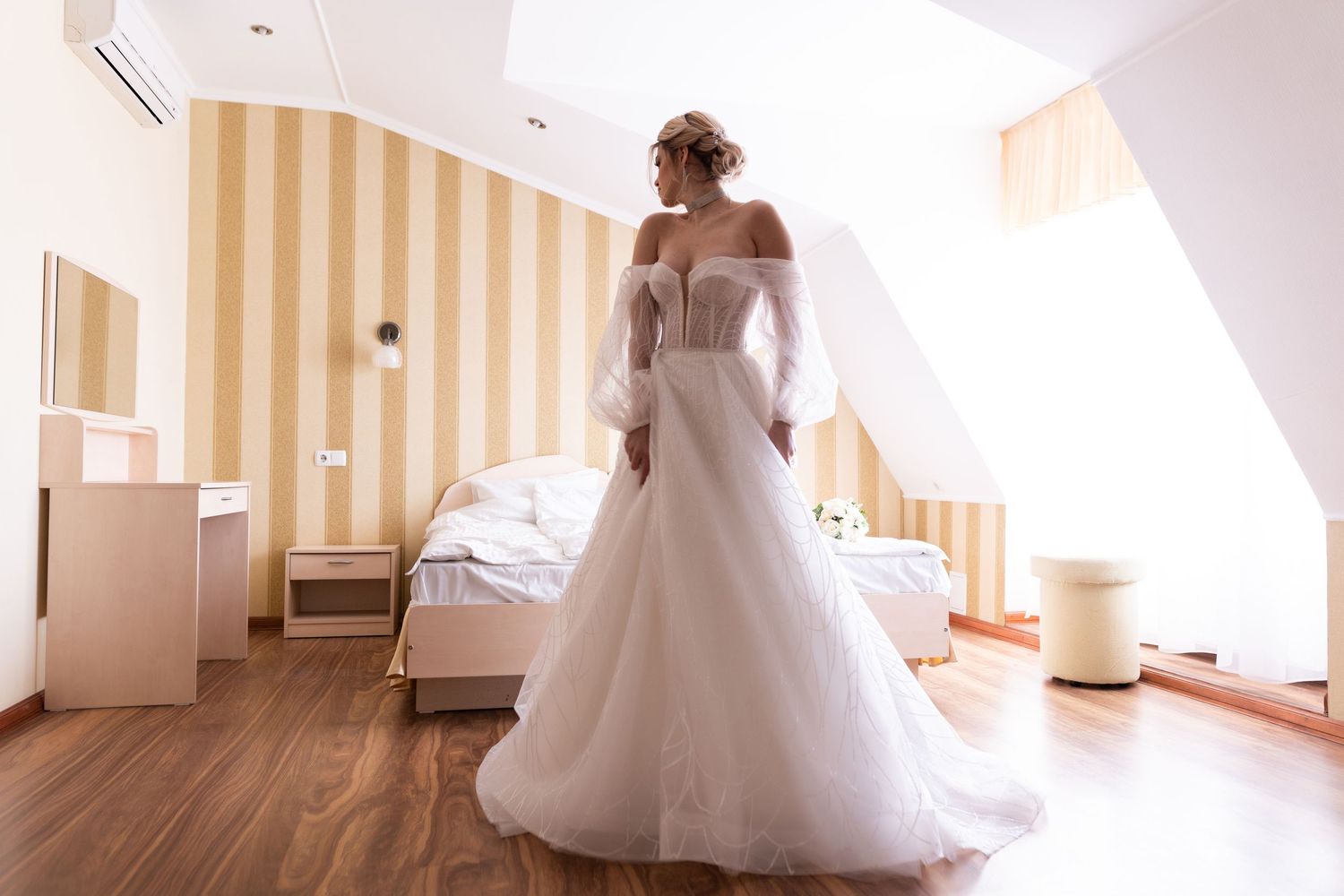 A bride in a flowing white wedding gown stands in a sunlit hallway with striped wallpaper.