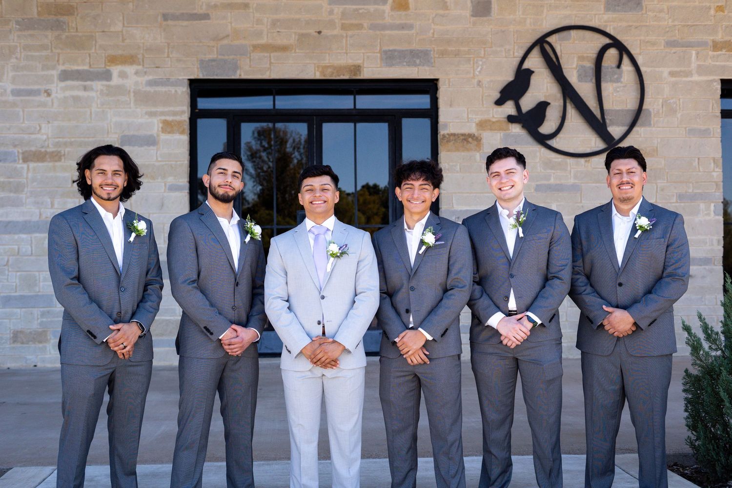 Wedding party members in gray suits and one in light blue stand together against a brick wall outdoors.