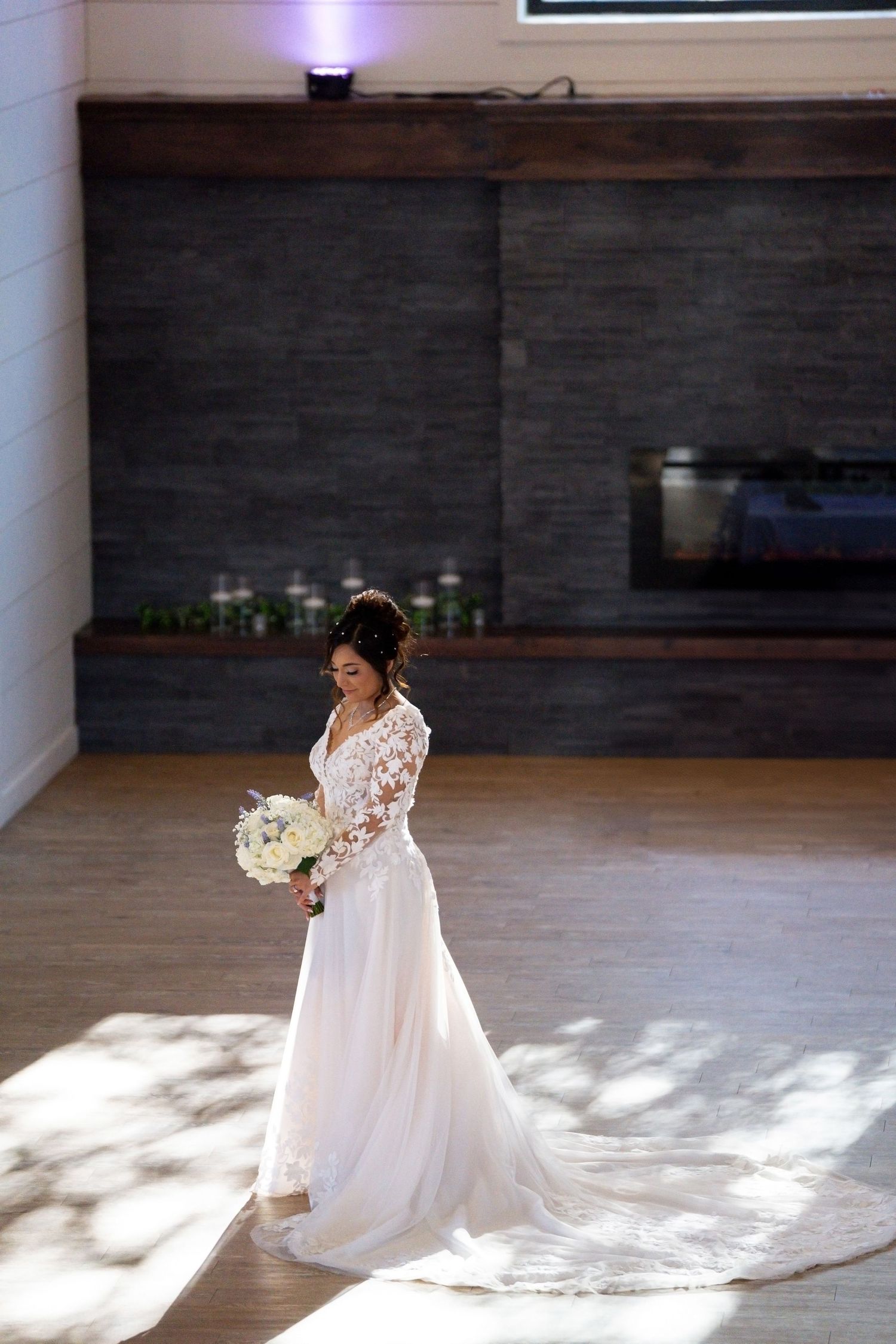 Bride in flowing white gown with long train stands in sunlit indoor space.