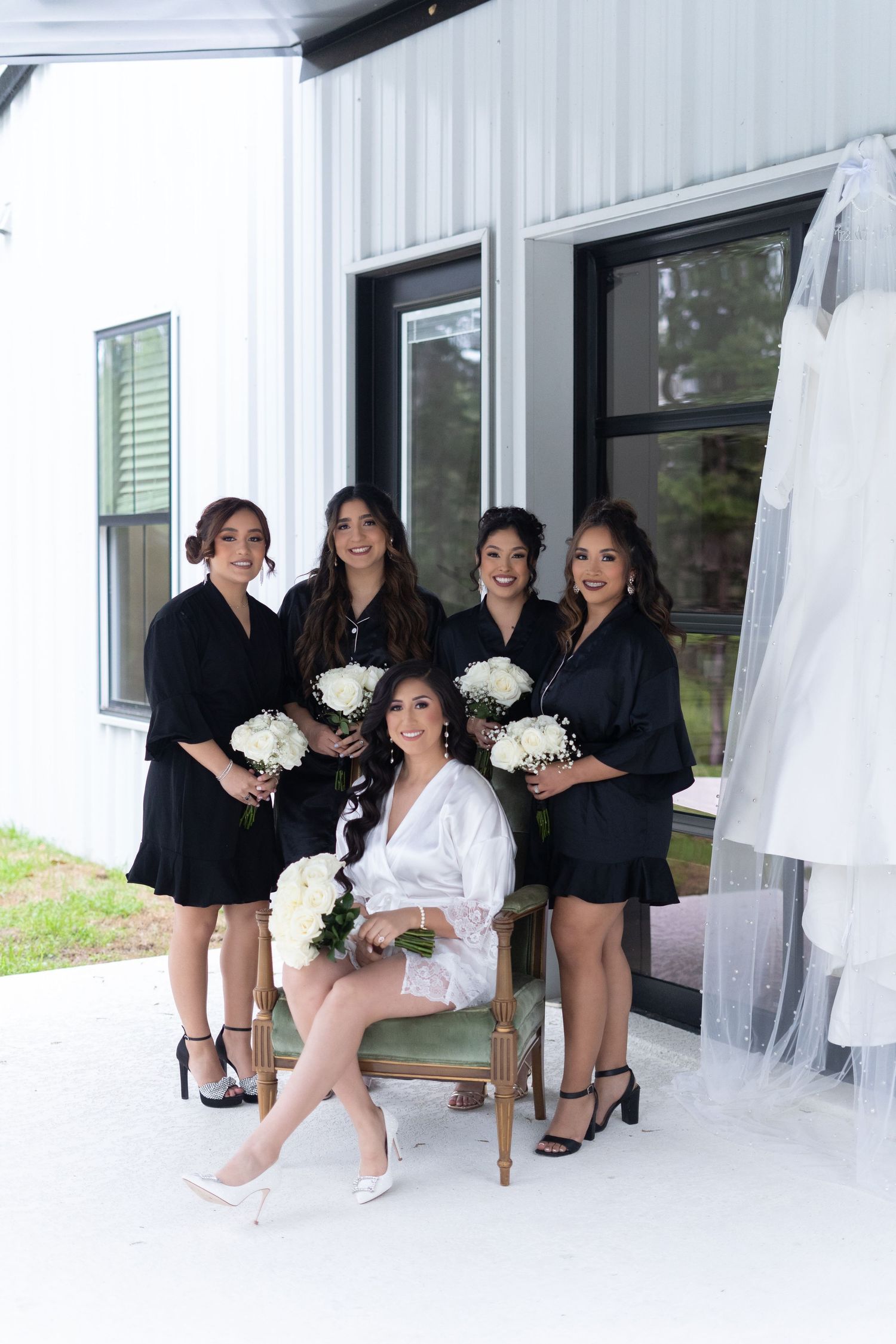 Bridesmaids in black robes pose together on white chairs against a white wall holding matching bouquets.