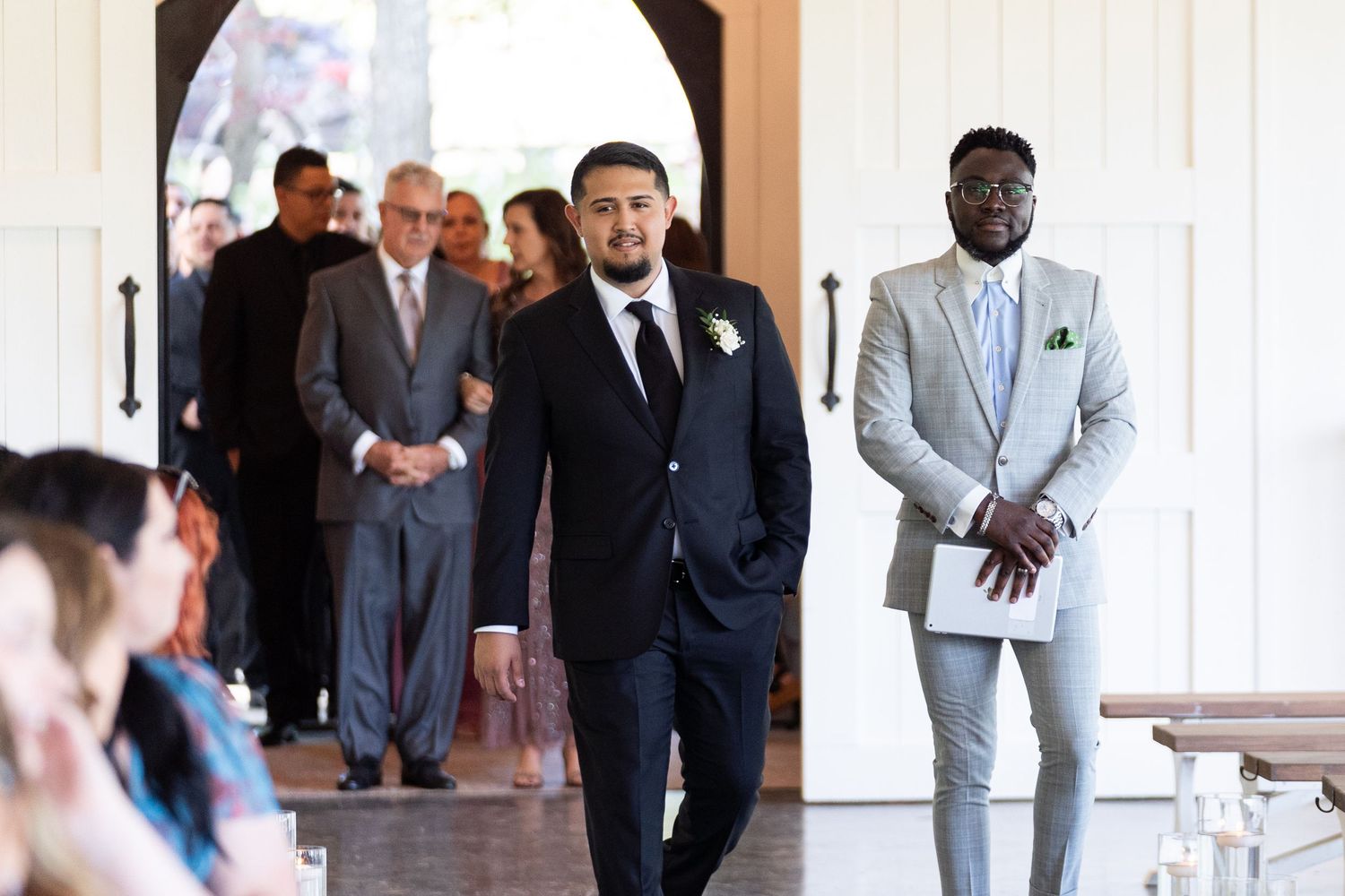 Groomsmen in formal suits stand together in a bright hallway during a wedding celebration.