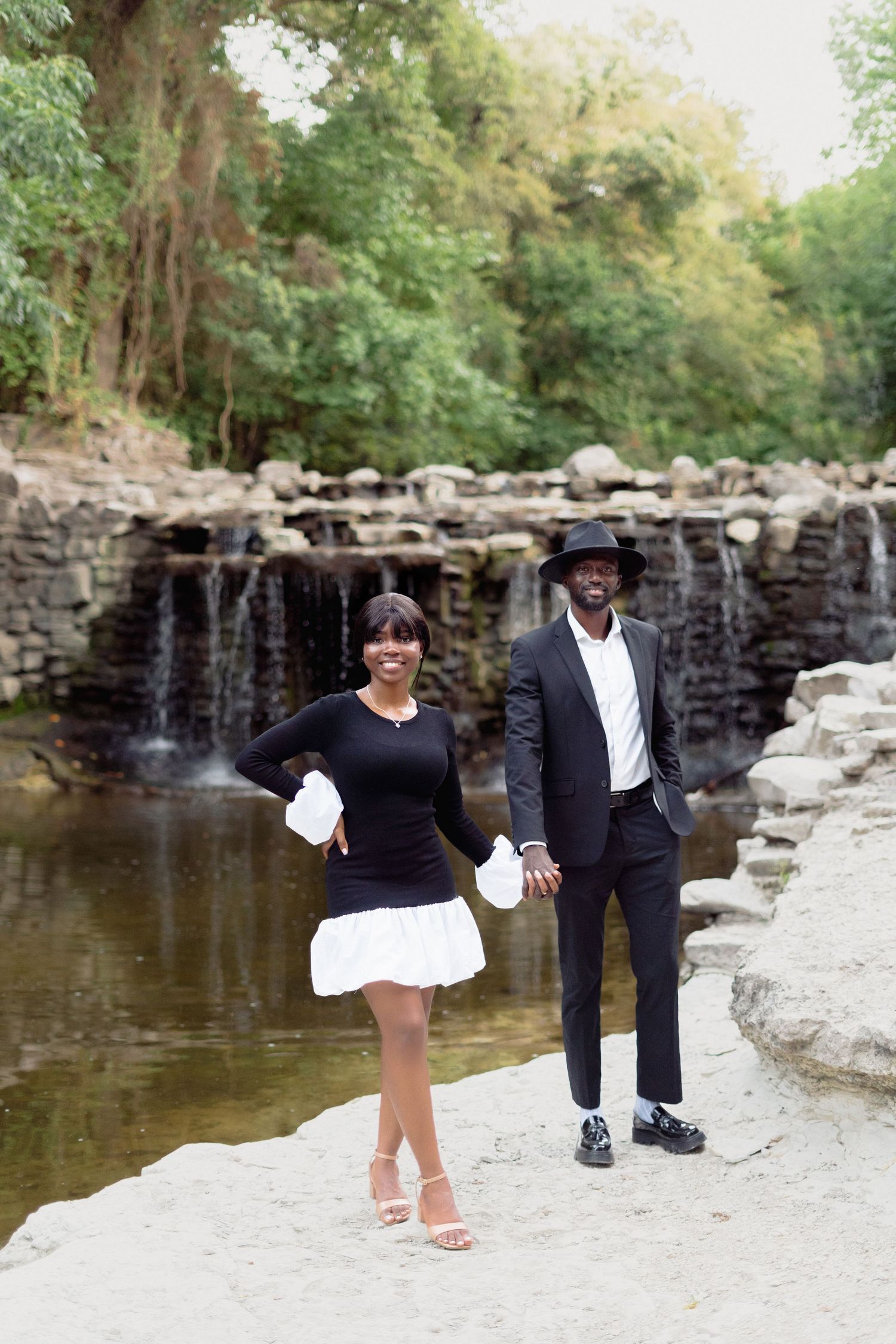 A couple poses by a waterfall, wearing contrasting black and white outfits in an outdoor setting.