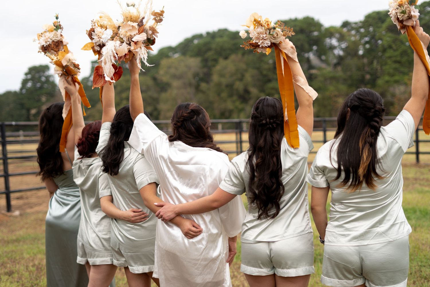 Wedding party in matching sage green dresses pose with raised arms in outdoor setting.