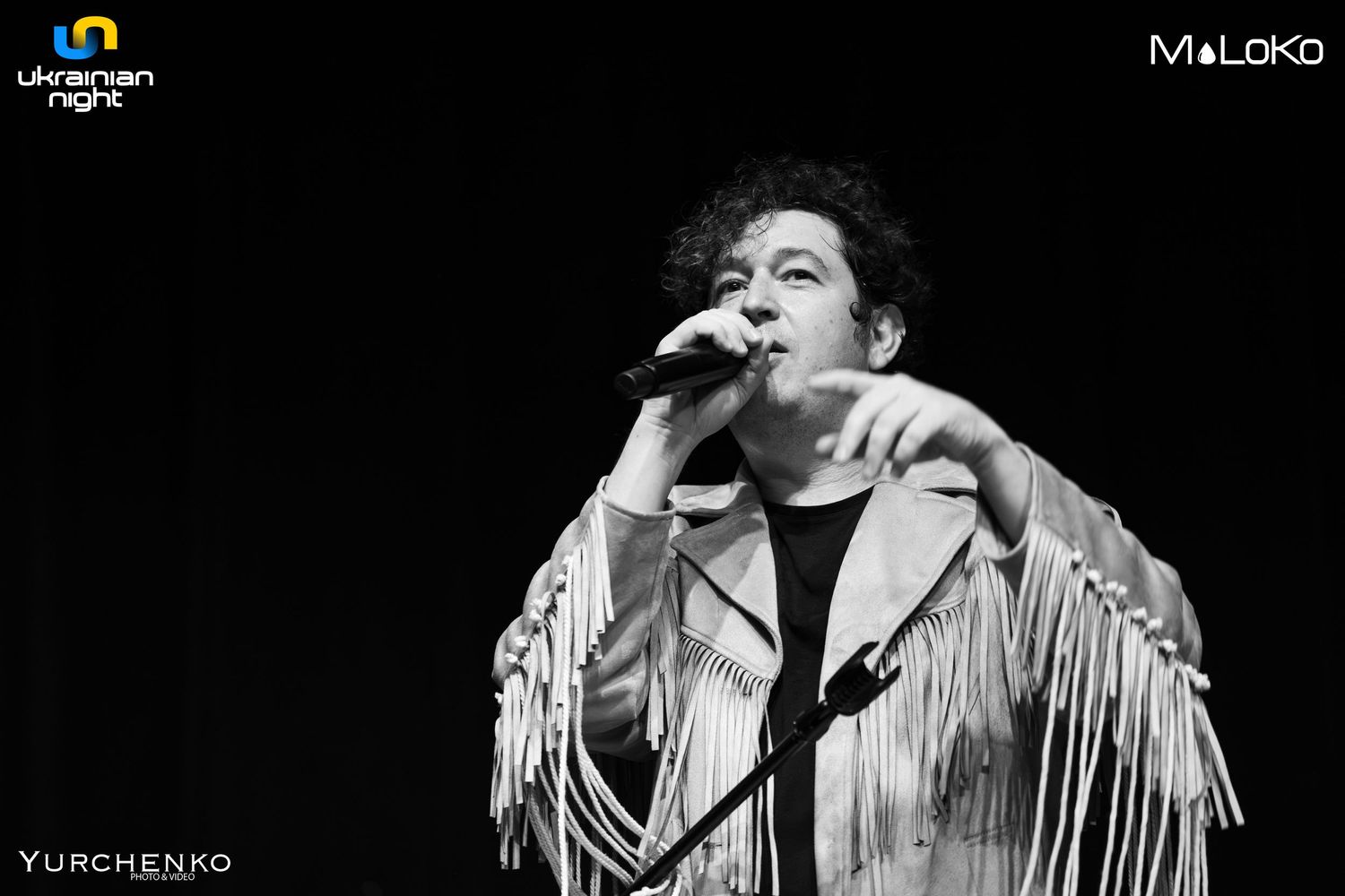 Performer in fringed jacket gesturing dramatically on dark stage with moody lighting.