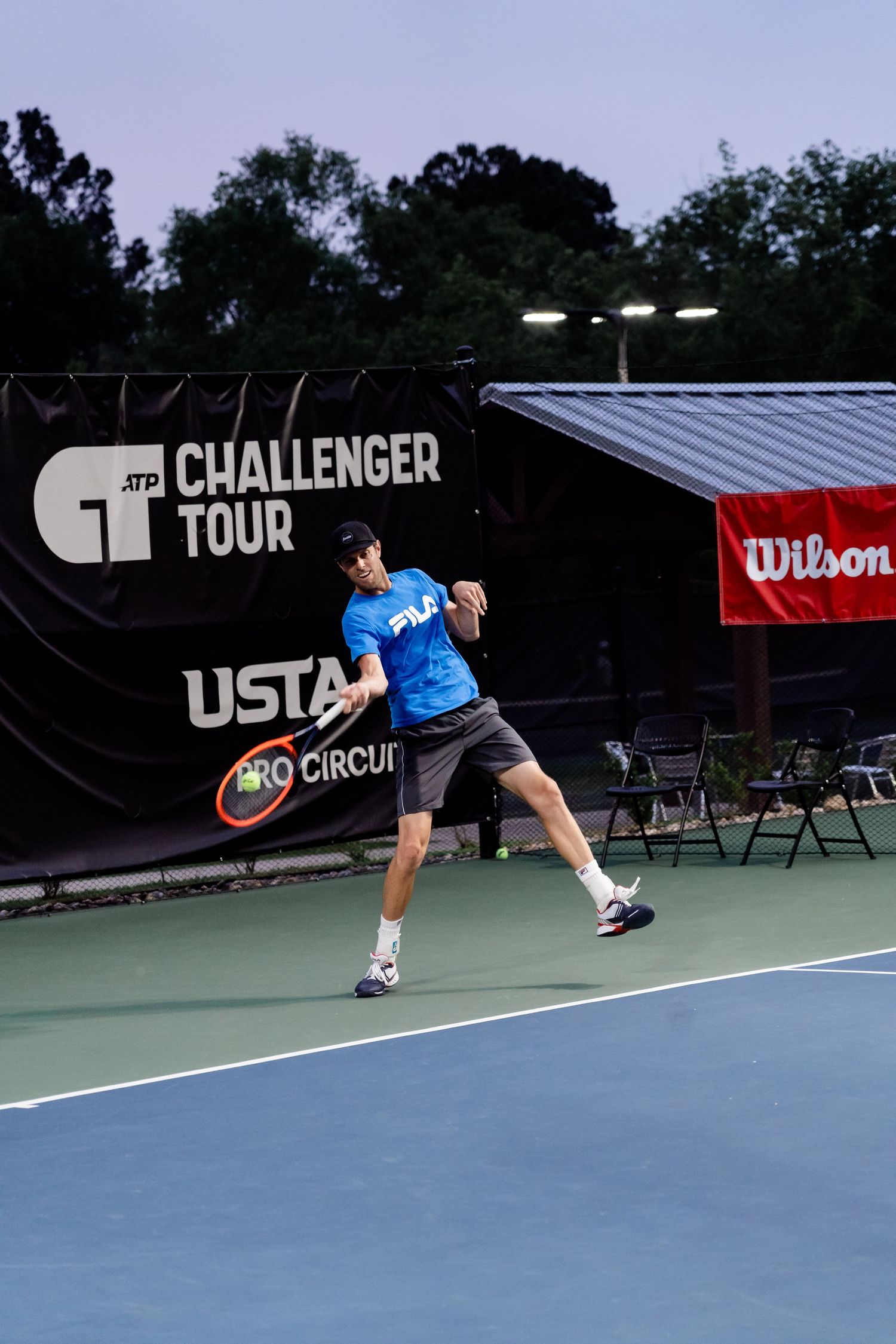 Tennis player in blue shirt stretches for a shot on outdoor court during USTA Challenger tournament.